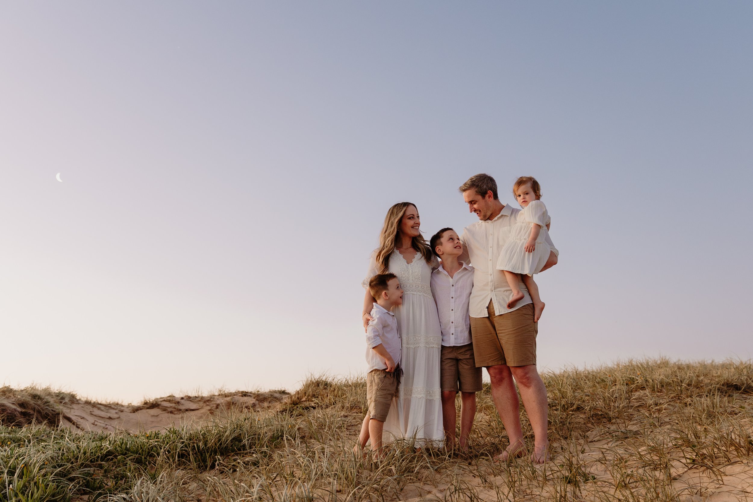 A family of five standing on sand dunes at sunset, the parents holding two children, with a clear sky and a crescent moon in the background.