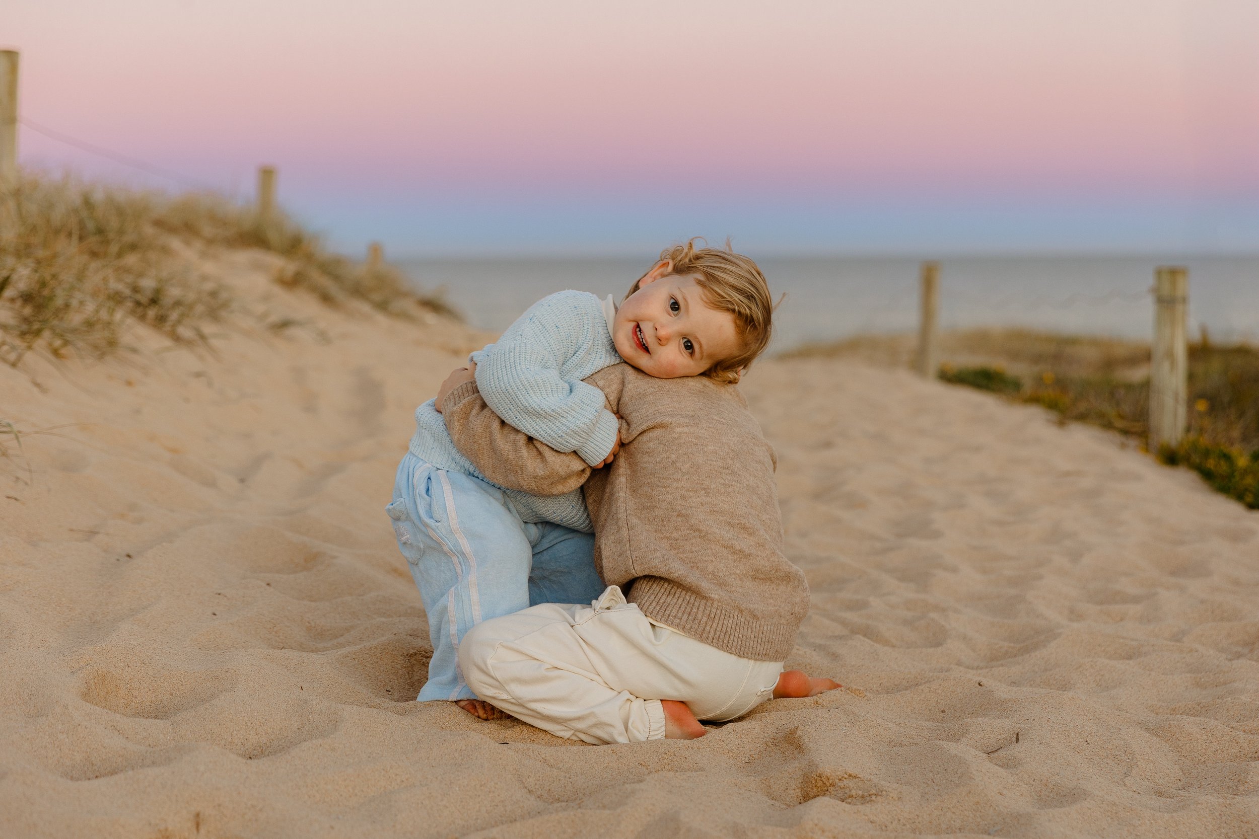Two children, a boy and a girl, hugging on a sandy beach at sunset, with pastel pink and blue sky in the background.