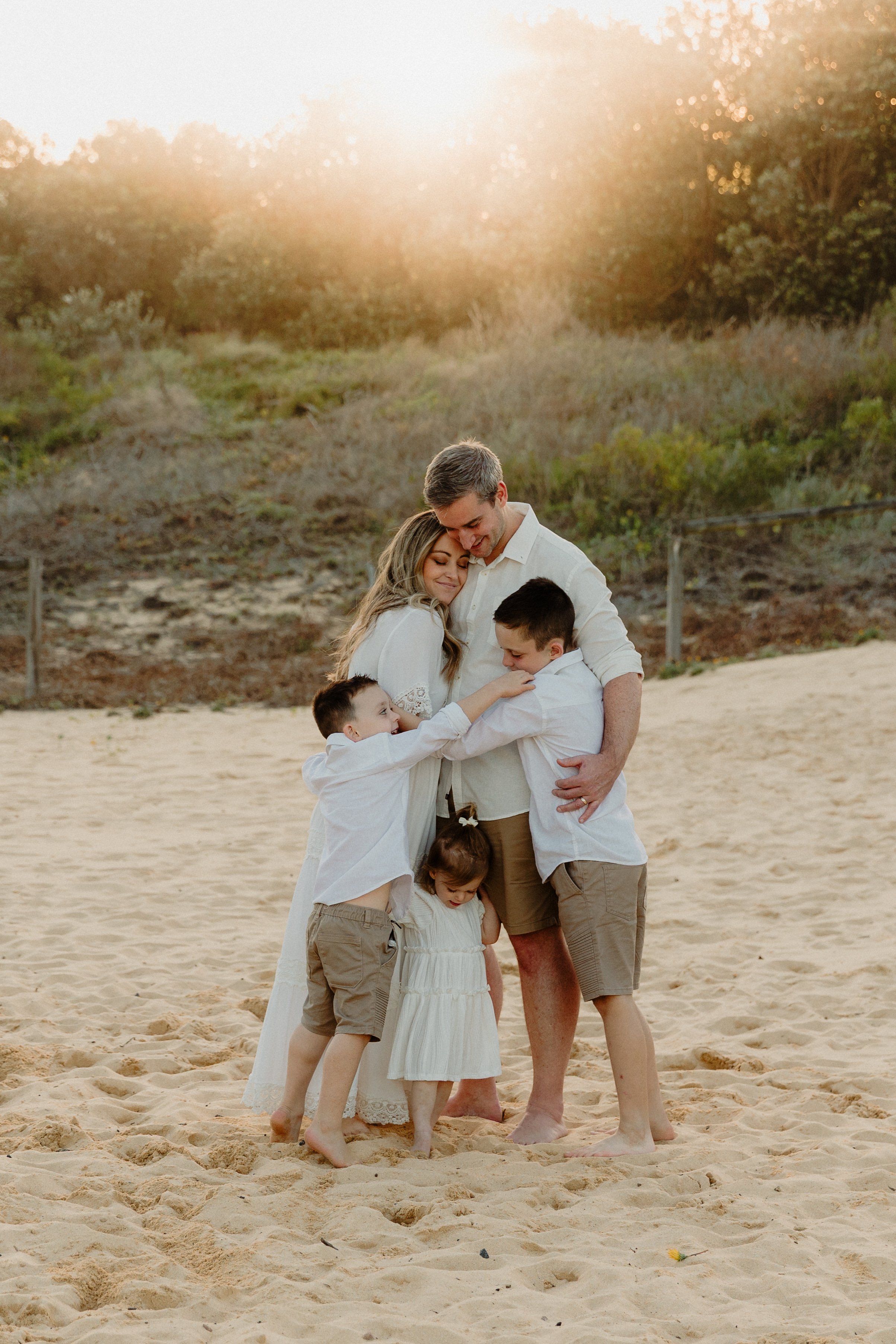 A family of five hugging and smiling on a sandy beach at sunset, with trees in the background.