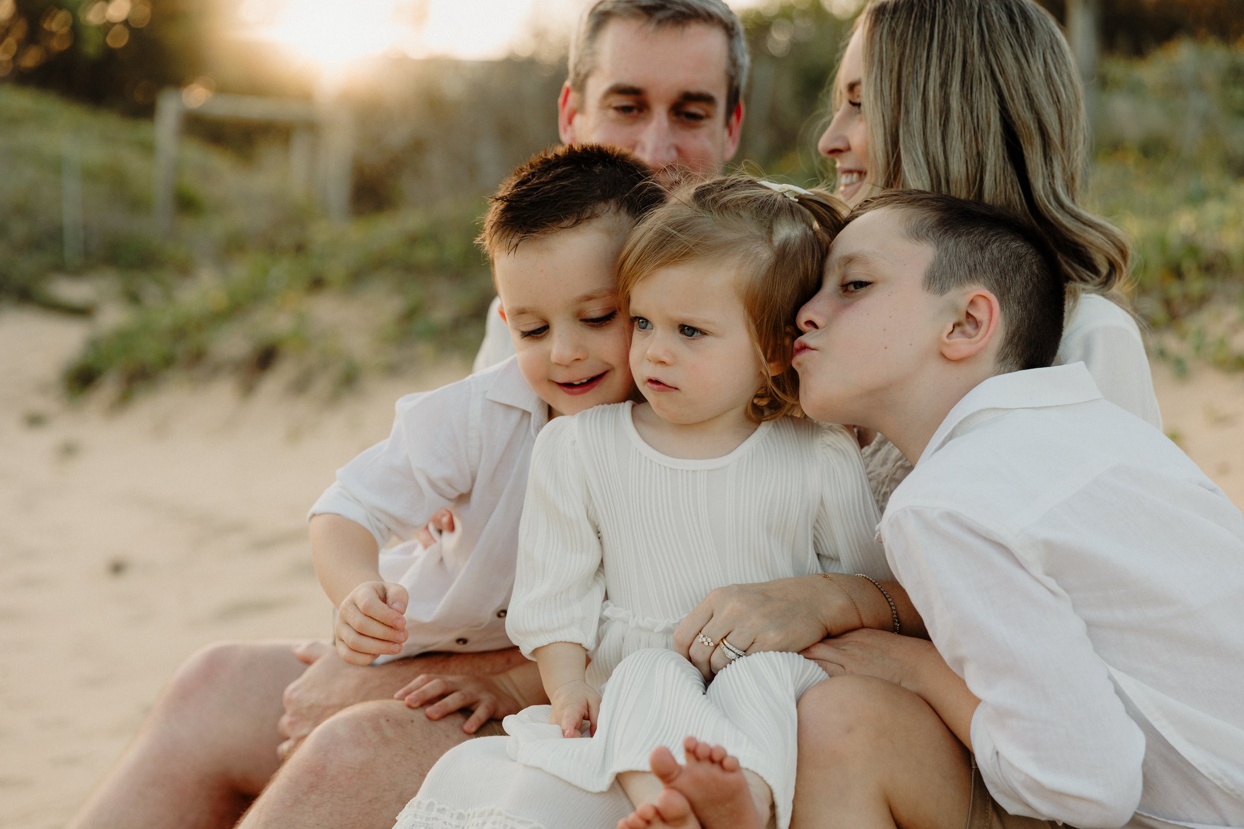 A family of six, including three children and two adults, sitting closely together outdoors on a beach at sunset, all dressed in white and looking at something with curiosity.