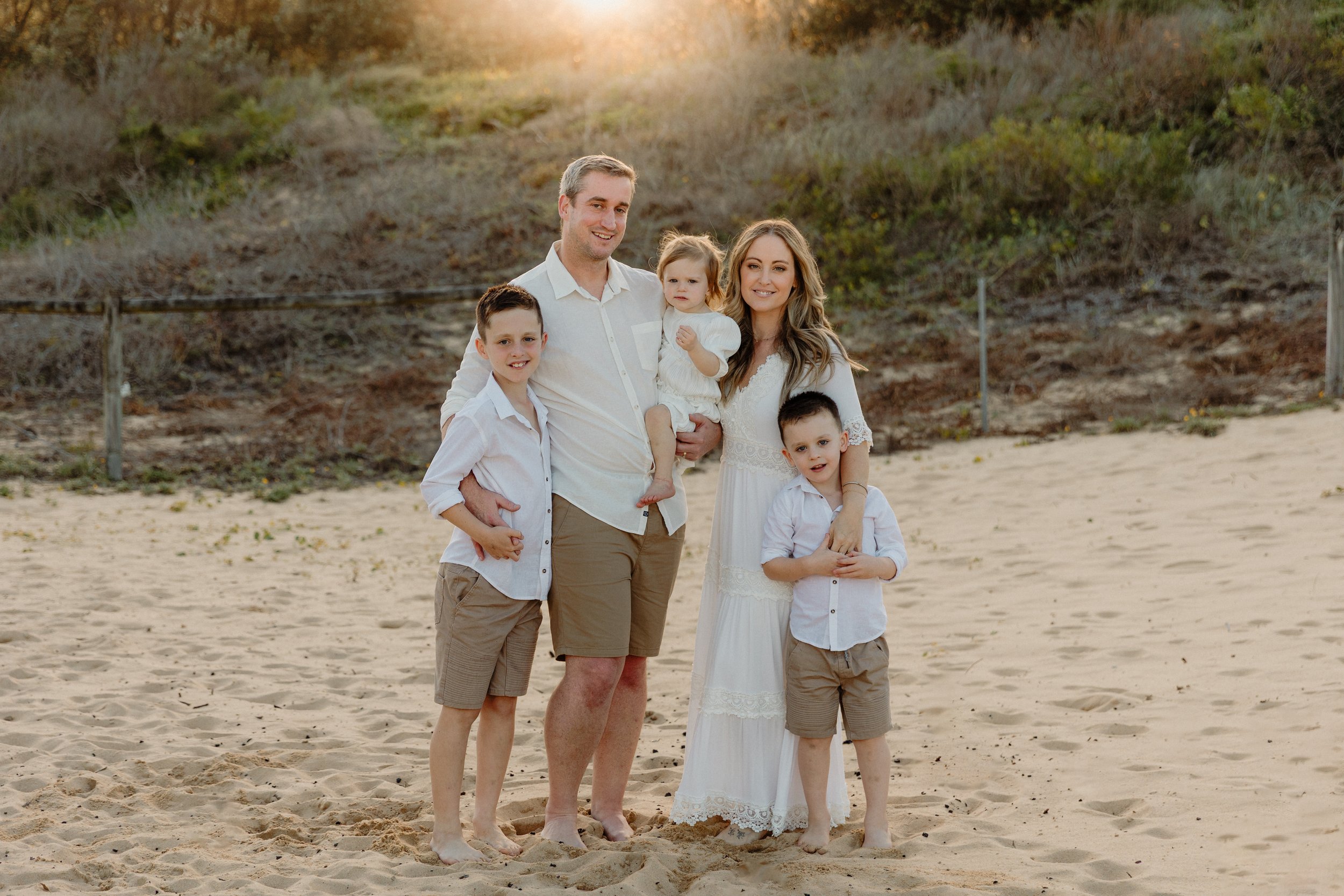 A family of six standing on a sandy beach at sunset, smiling at the camera, dressed in white and beige clothing.