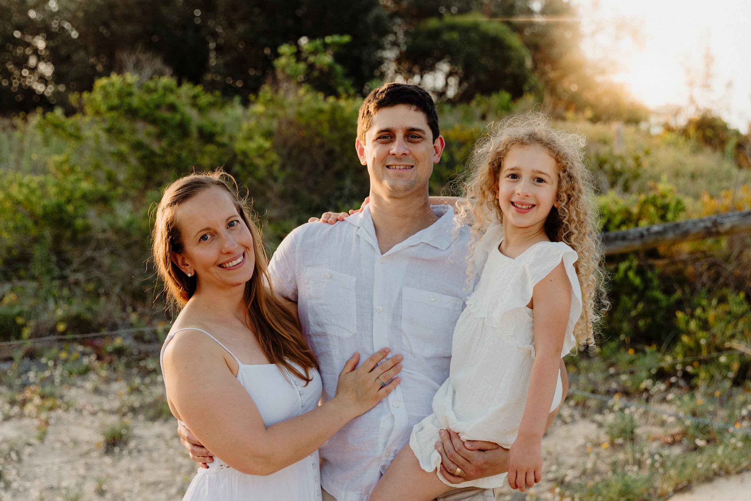 A family of three standing outdoors during sunset, smiling, with greenery and a fence in the background.