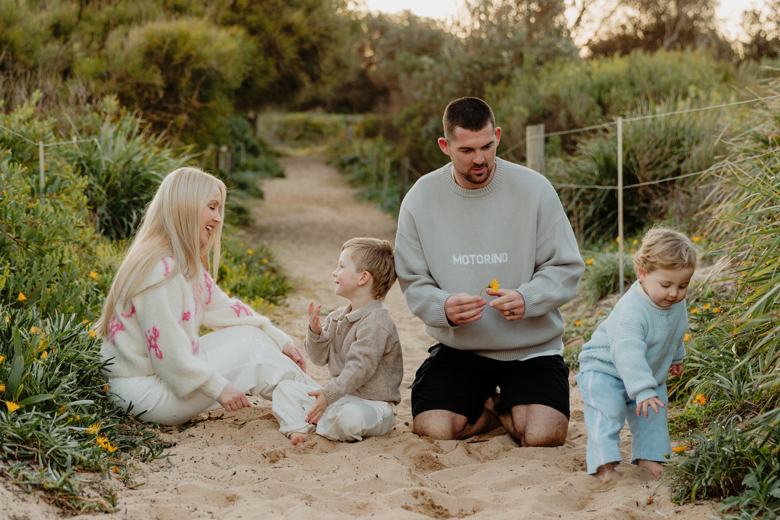 A family of five, including a blonde woman, two young boys, and two men, is enjoying a day on a sandy trail surrounded by green bushes and blooming flowers, with some members sitting and others kneeling in the sand.