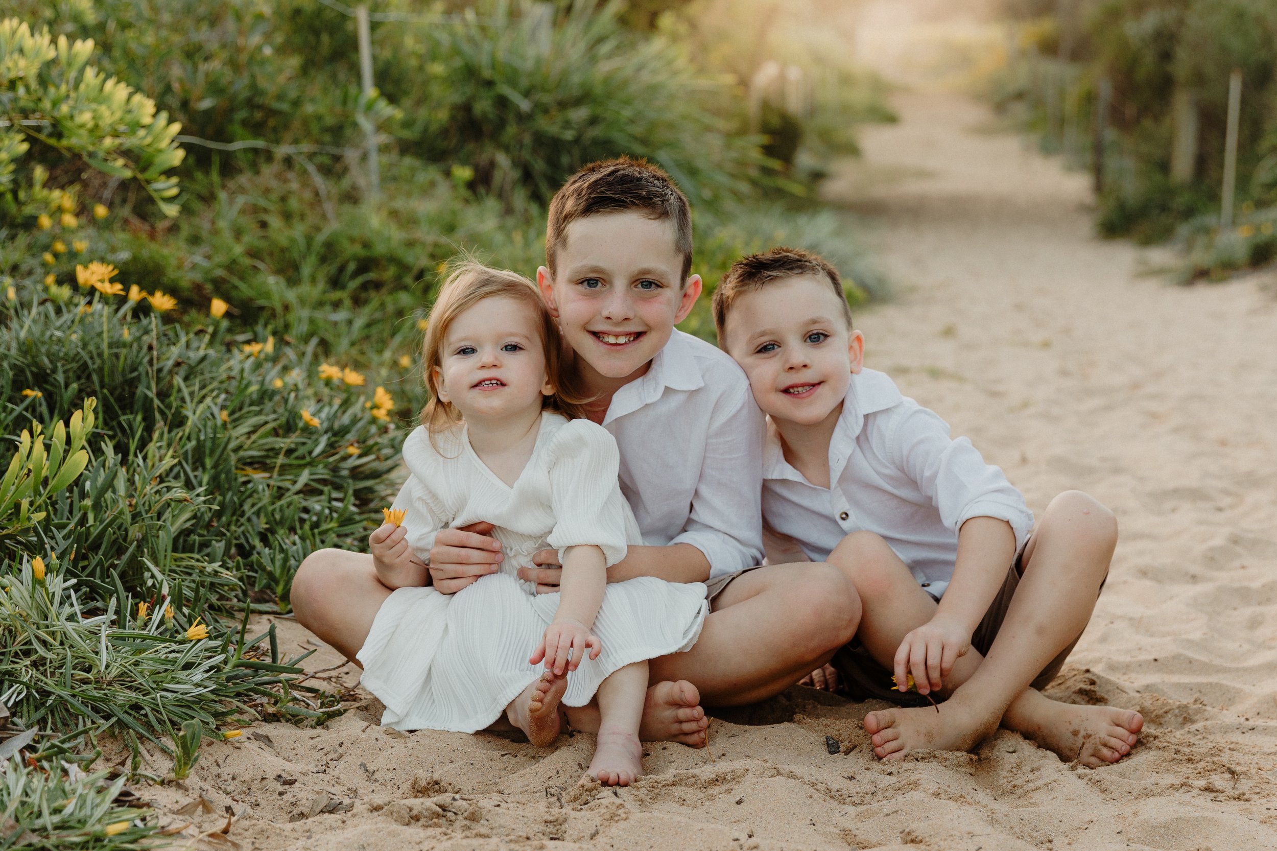 Three children sitting on sandy beach, smiling at camera. Green plants with yellow flowers on left, sandy path in background.
