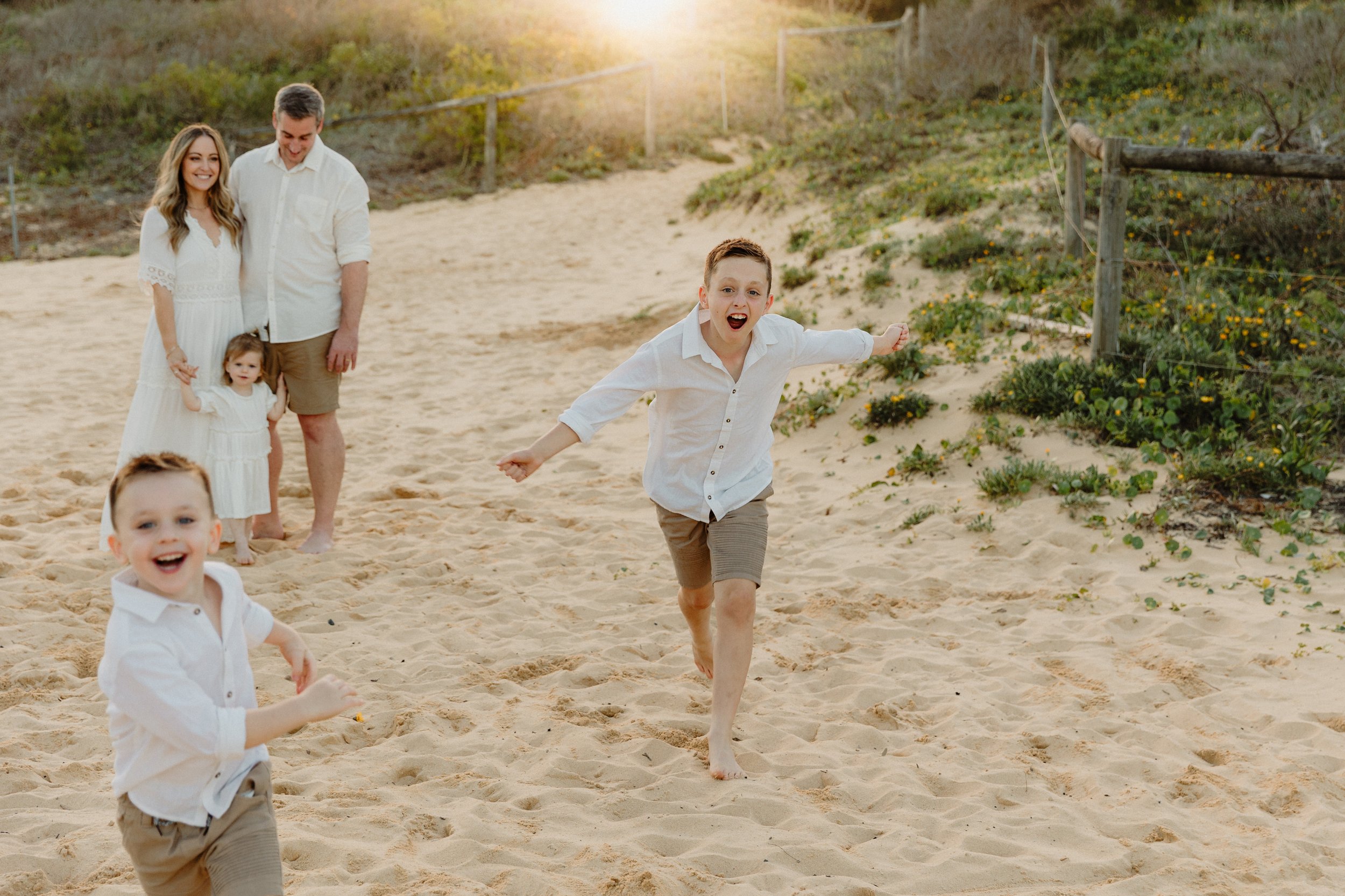 A family of five at the beach, including two young boys running and two girls, with an adult couple in the background, with greenery and a wooden fence on a sandy beach at sunset.