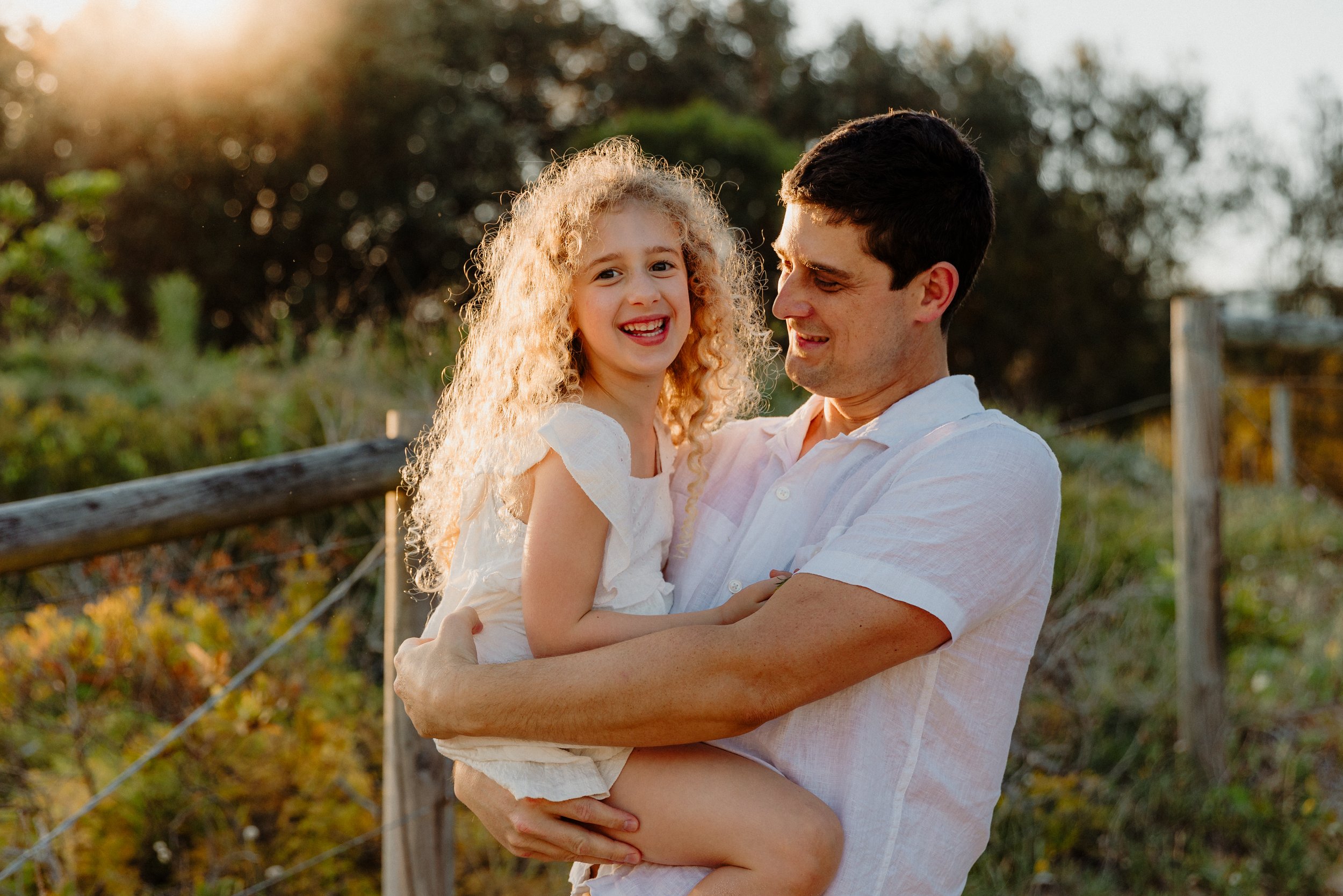 A man holding a young girl outdoors during sunset, both smiling and wearing white clothing.