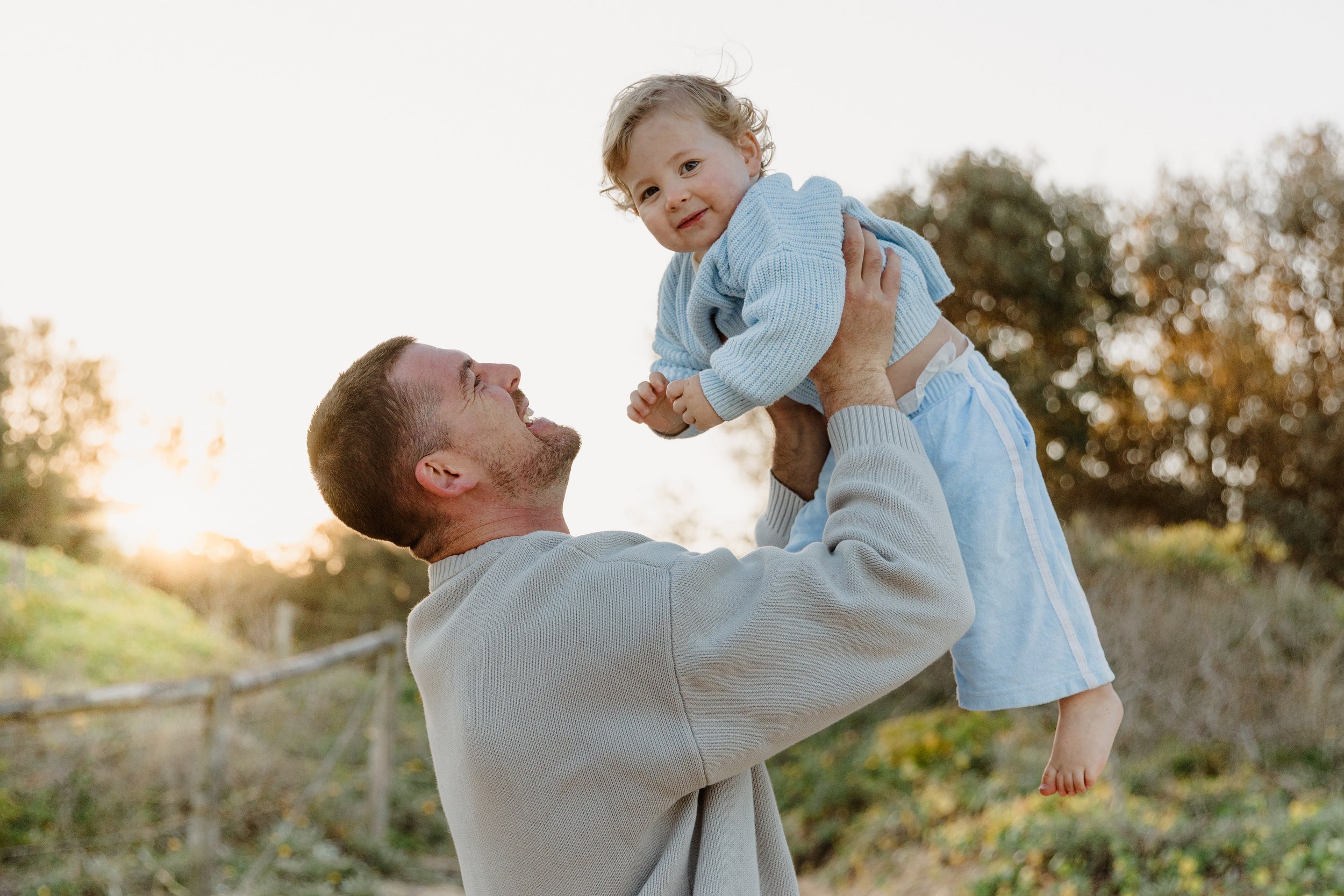 A man lifting a young child in the air outdoors at sunset, both smiling and enjoying a moment together in a natural setting.