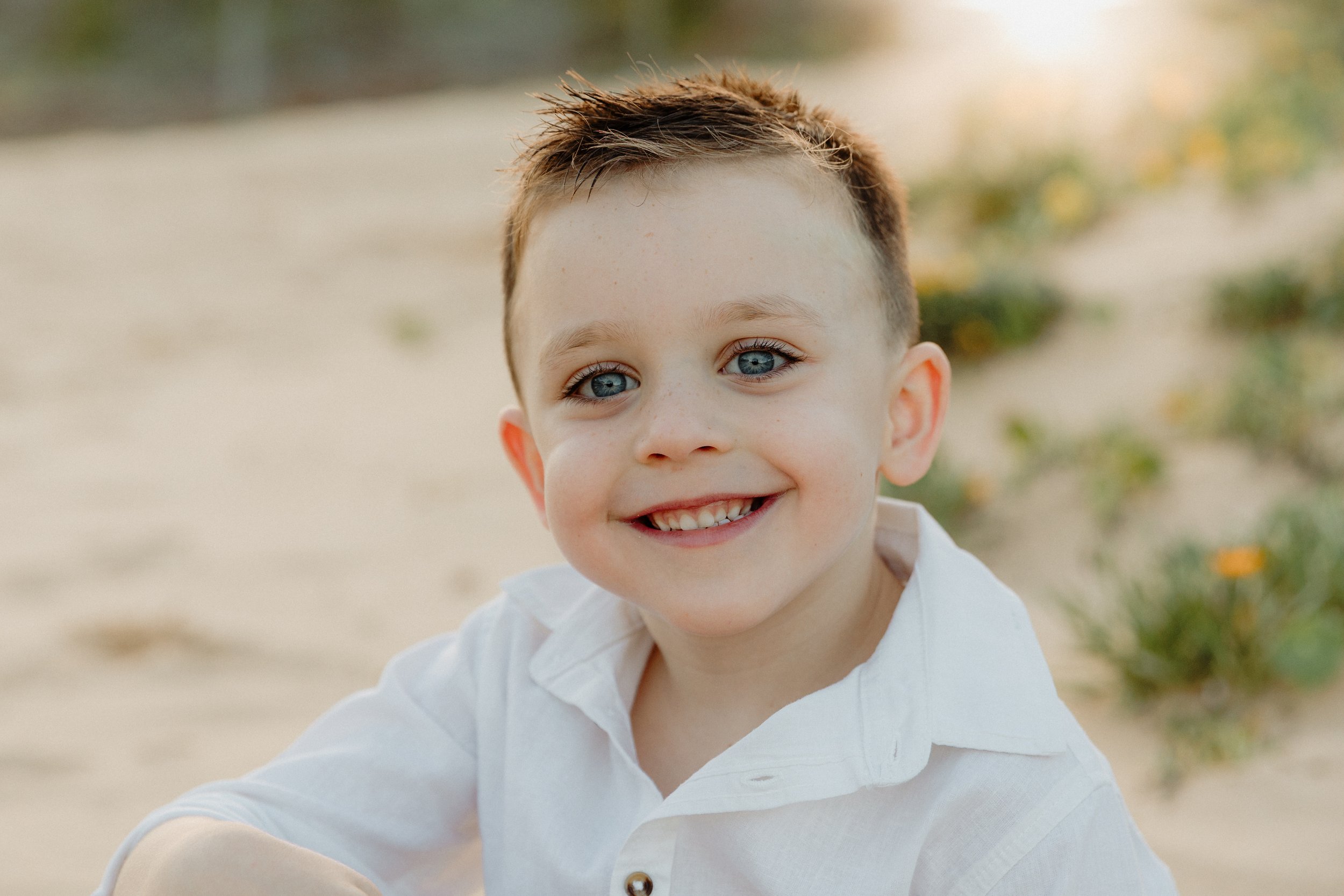 Close-up of a smiling young boy with blue eyes and brown hair, wearing a white shirt, sitting outdoors on a sandy area with some greenery and yellow flowers in the background.