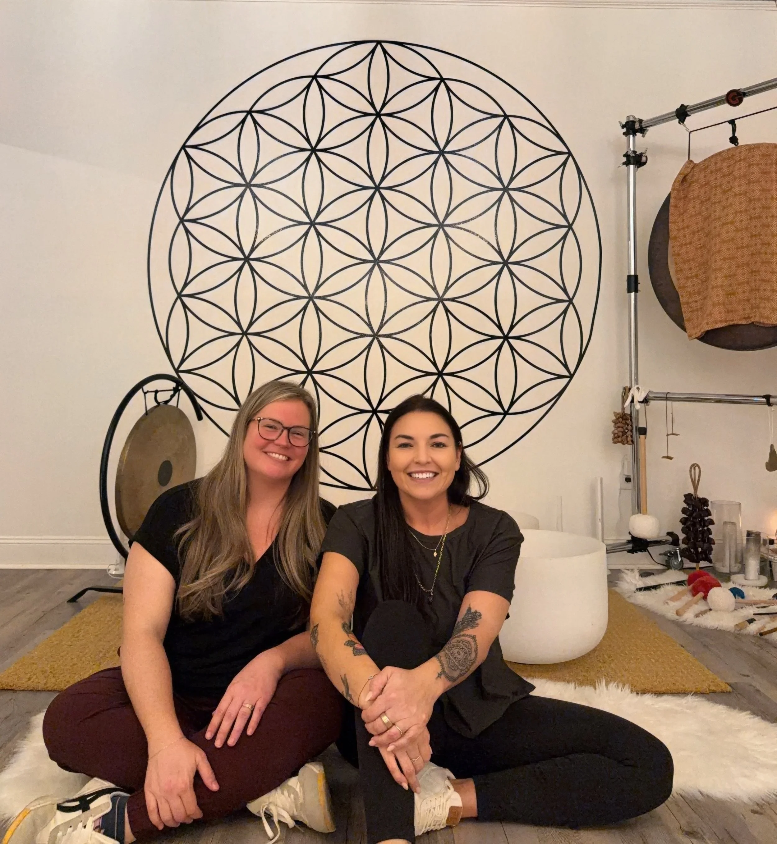 Two women sitting on the floor in front of a wall with a large geometric circular wall art. One woman has long blonde hair and glasses, the other has long dark hair and tattoos on her arms. They are smiling.
