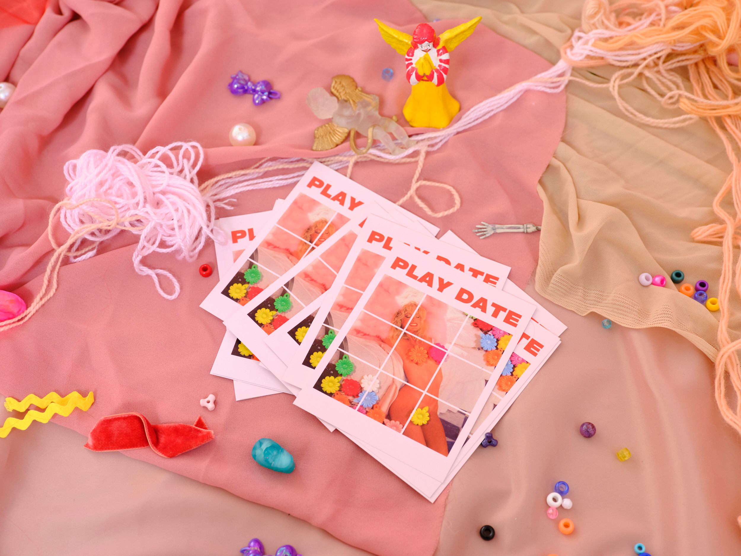 Pink and beige fabric with scattered decorative items such as a small angel figurine, colorful beads, paper crafts labeled 'PLAY DATE' and 'PLAY', yarn, and small plastic accessories.