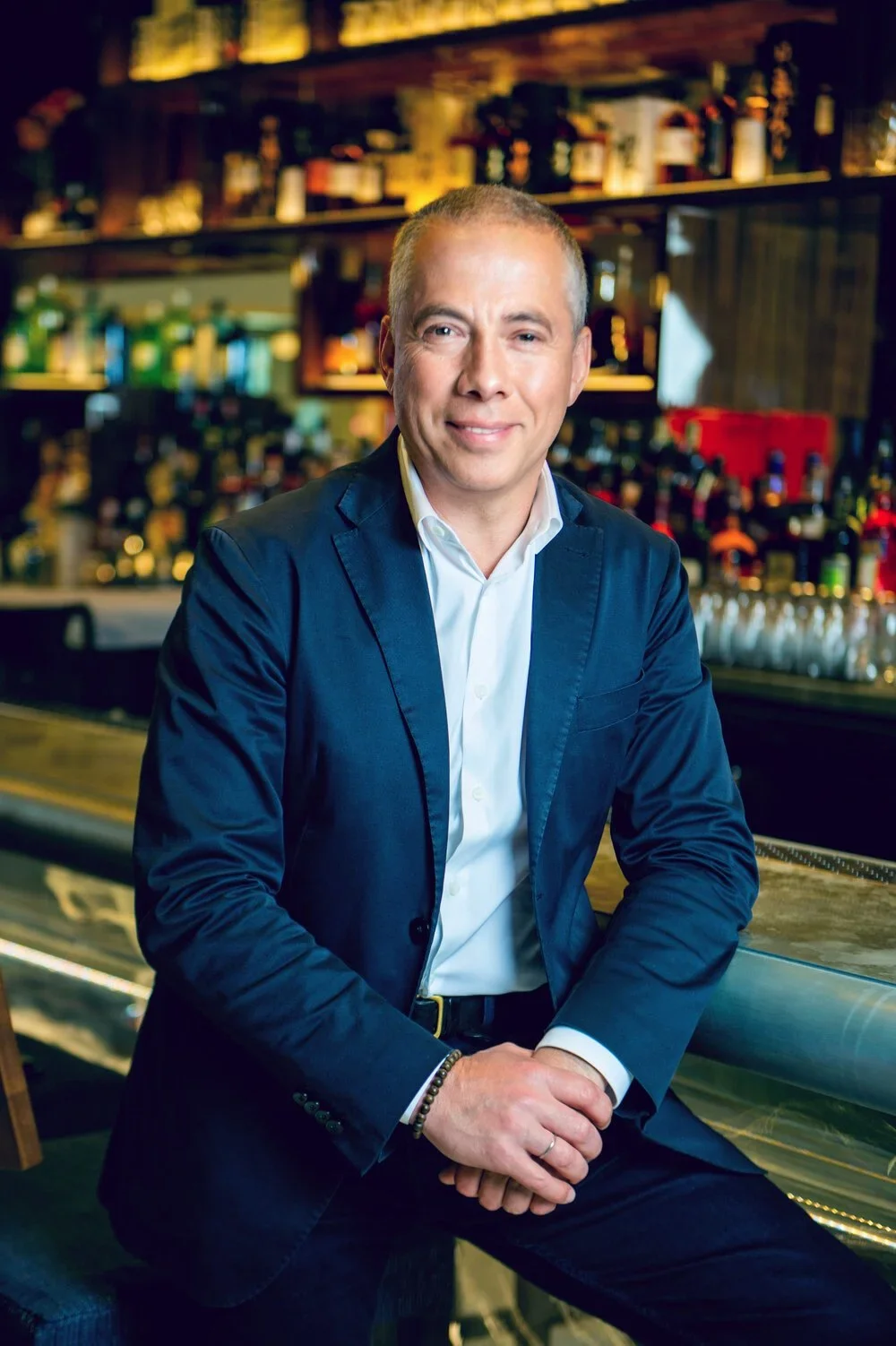 A man in a navy blazer and white shirt sitting at a bar, smiling, with a backdrop of liquor bottles on shelves.