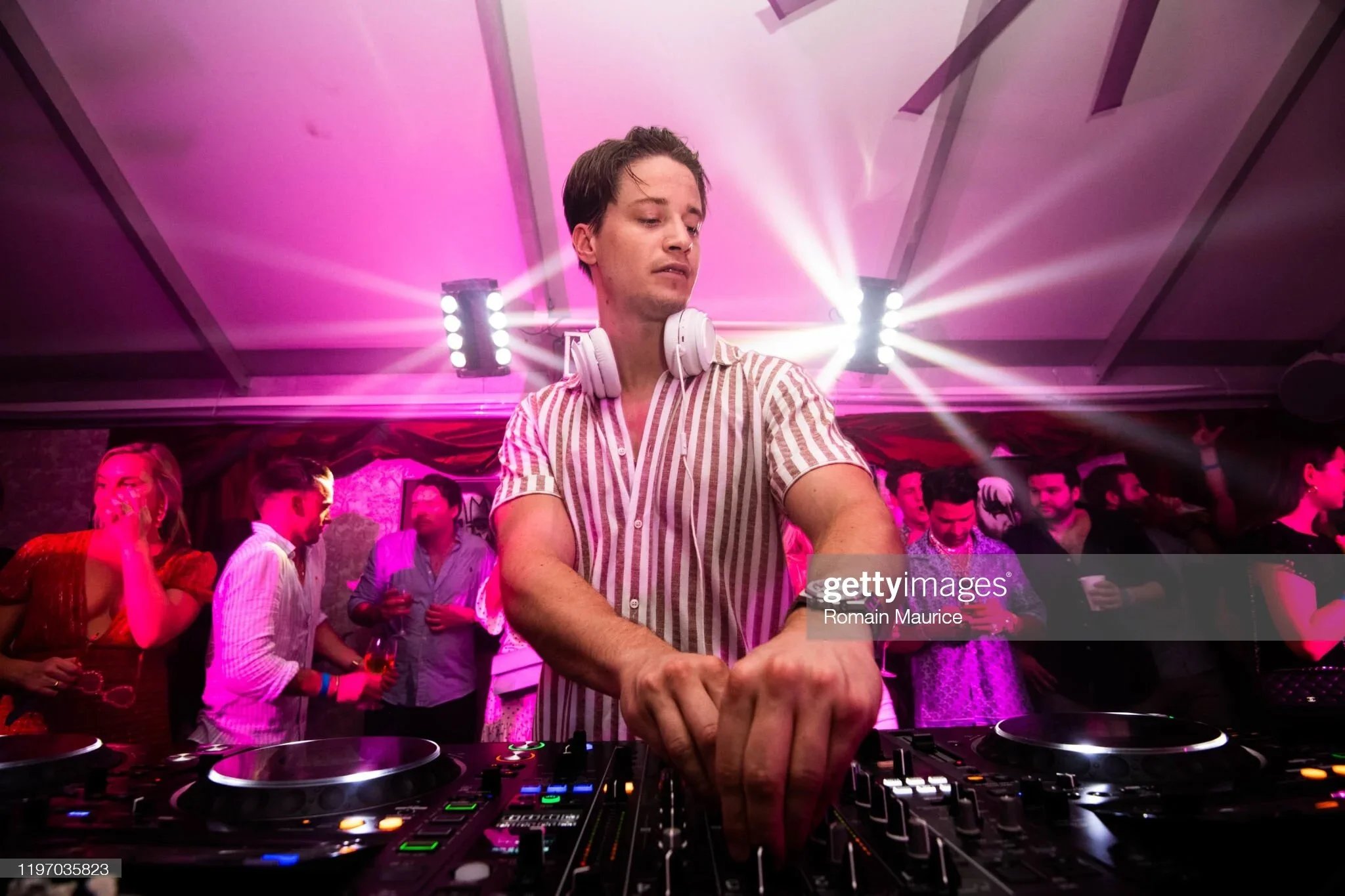 A DJ with headphones around his neck is performing at a nightclub with pink and purple lighting. People are dancing and socializing in the background.