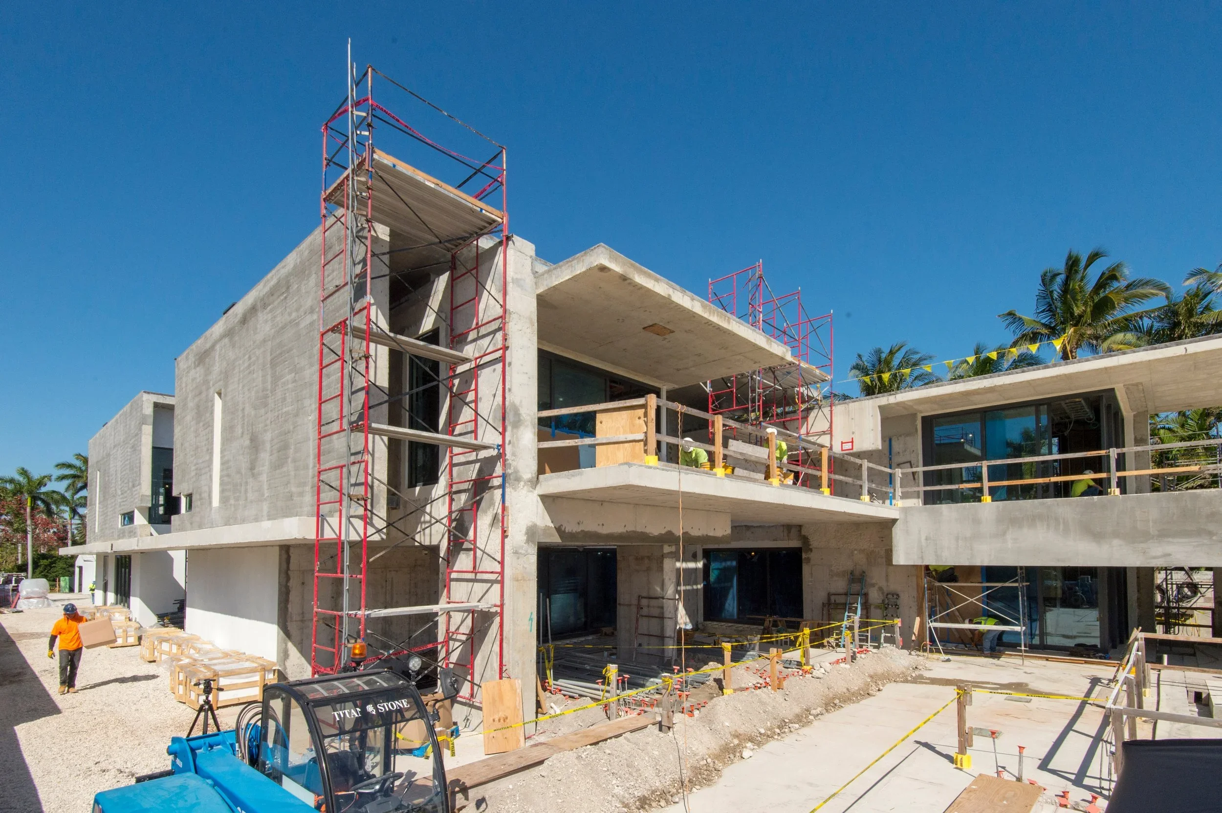 Multi-story building under construction with scaffolding, construction workers, and building materials, set against a clear blue sky and surrounded by palm trees.
