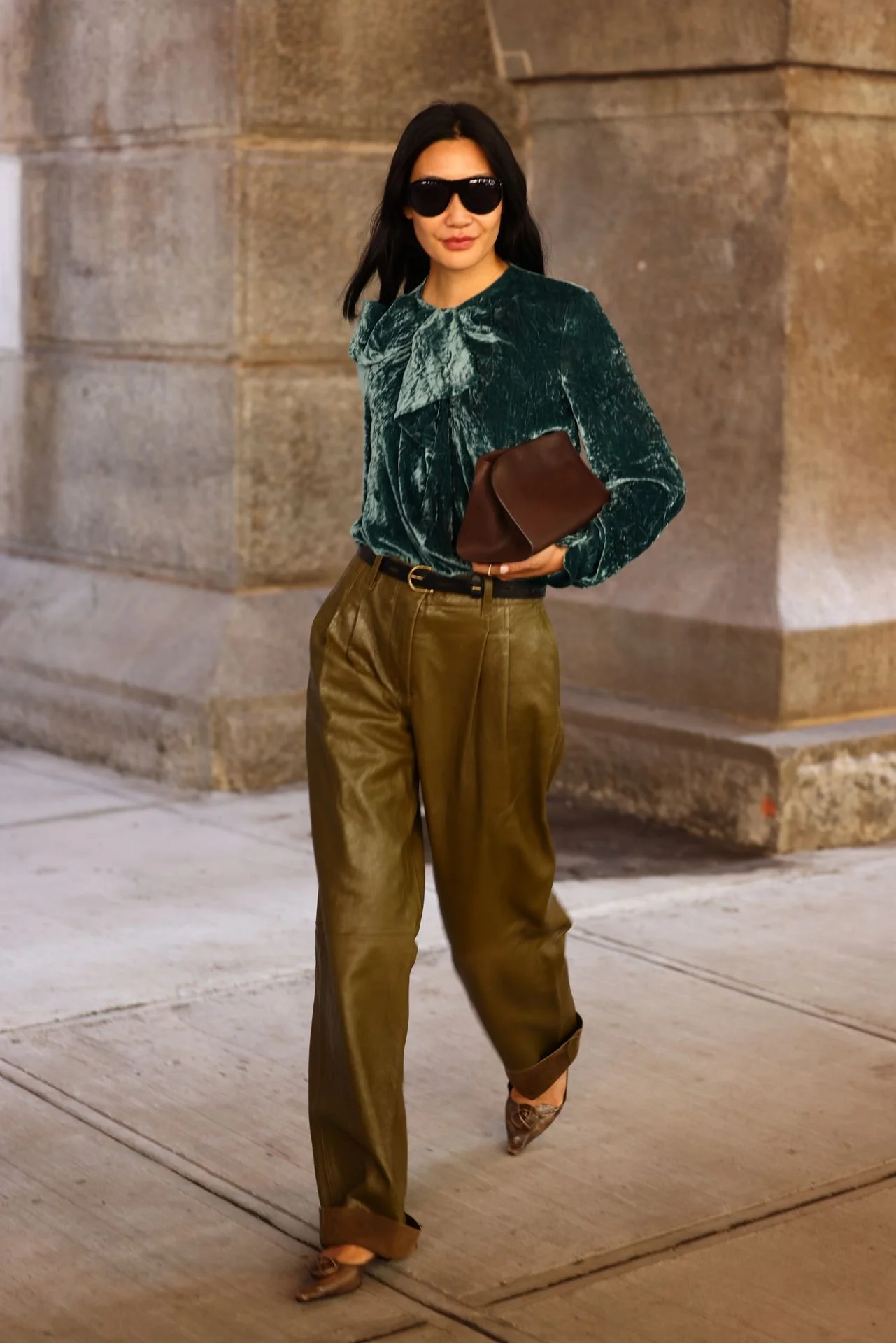 A woman with dark hair wearing black sunglasses, a velvet green top with a bow tie detail, loose brown leather pants, and high-heeled shoes walking on a city sidewalk at night.