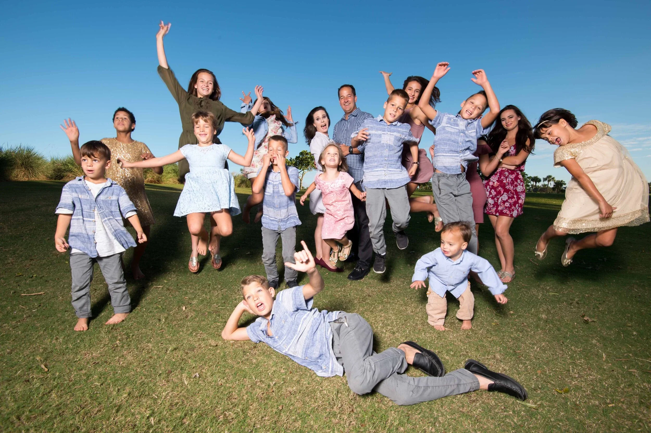 A group of children and adults outside on a sunny day, playing and smiling on a grassy field with trees and a clear blue sky in the background.