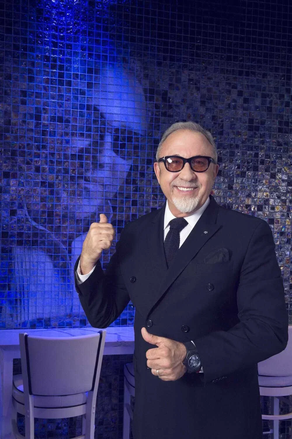 A smiling man wearing glasses, a black suit, and a black tie, standing in front of a blue mosaic wall with his thumb pointing towards his reflection or a mural of his face.