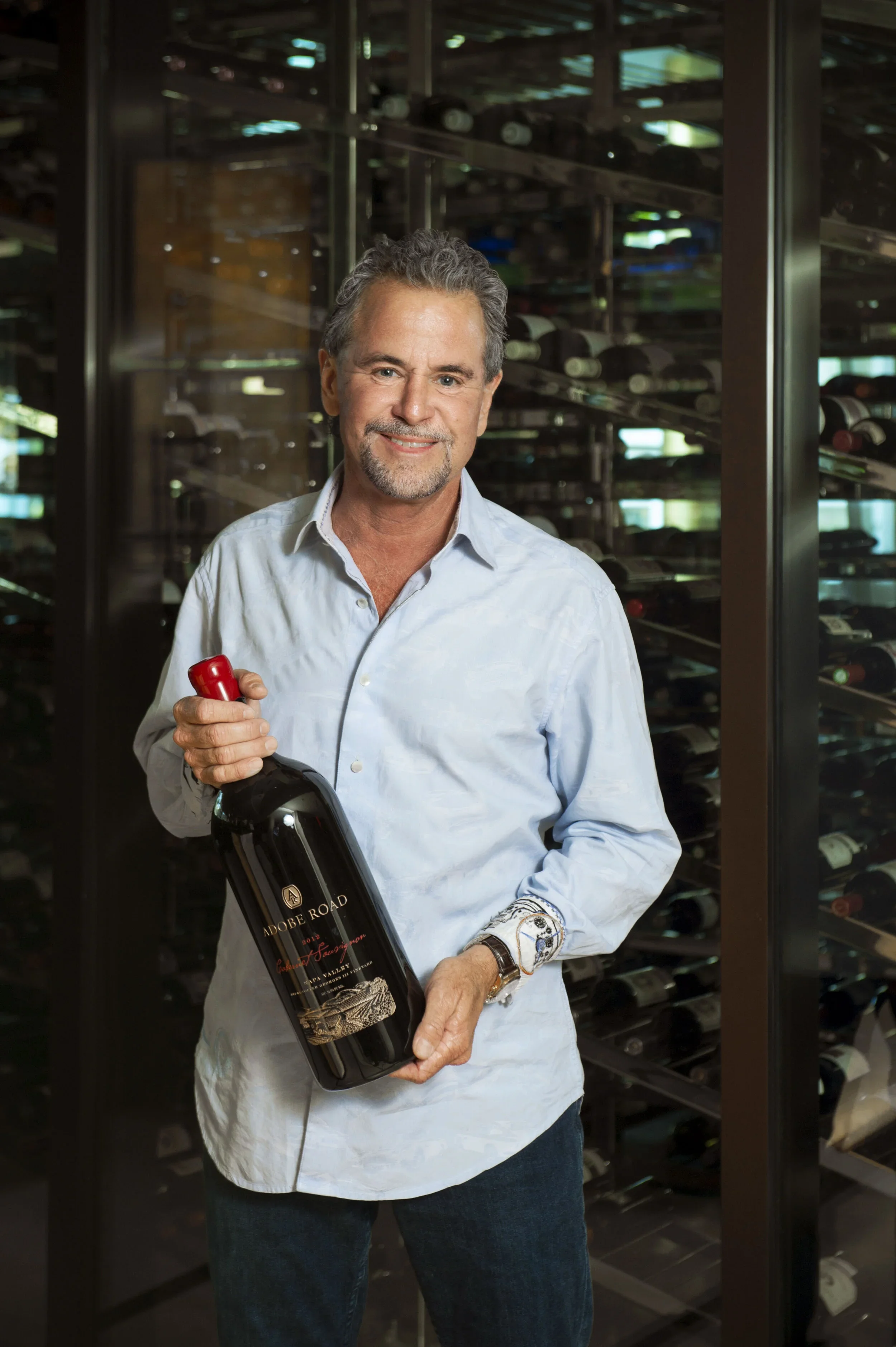 A middle-aged man with gray hair, beard, and mustache, smiling, holding a large bottle of wine labeled 'Adobe Road' in a wine cellar with bottles stored on metal racks in the background.