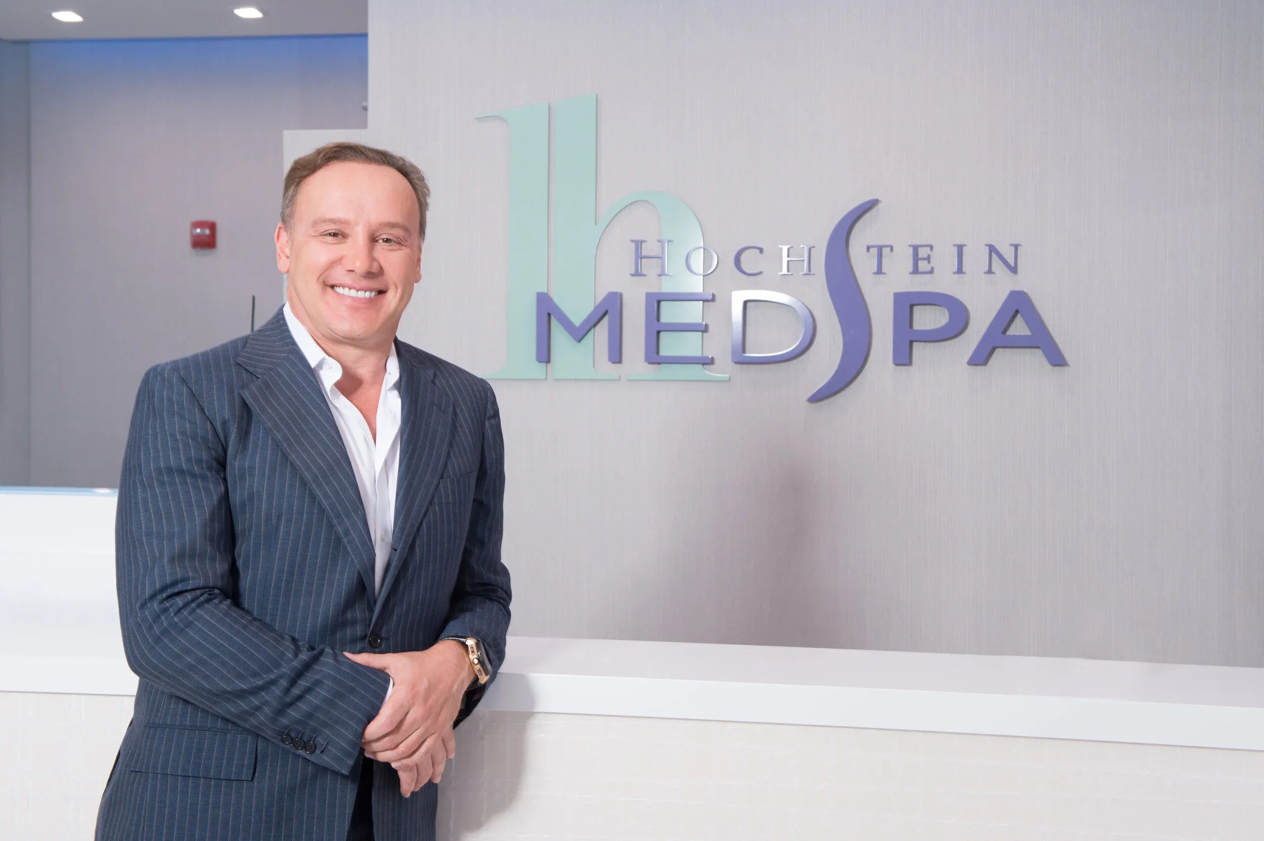 A man in a dark striped suit standing in front of a light-colored wall with a logo that reads 'Hochstein Media' and a large teal number '16' behind him, smiling at the camera.