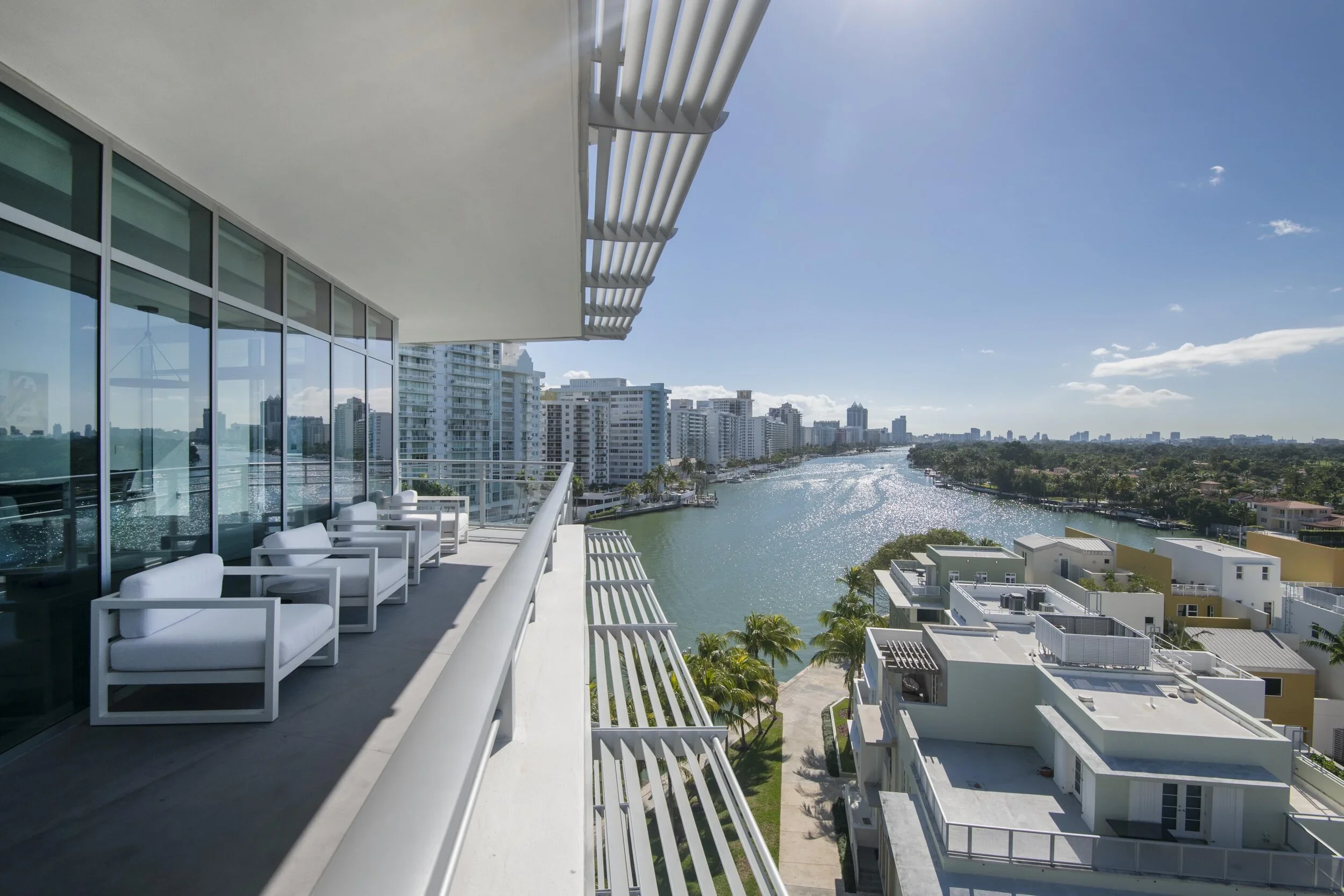 A balcony view from a modern high-rise apartment showing white outdoor furniture, glass sliding doors, and a cityscape with high-rise buildings along a waterway under a partly cloudy sky.