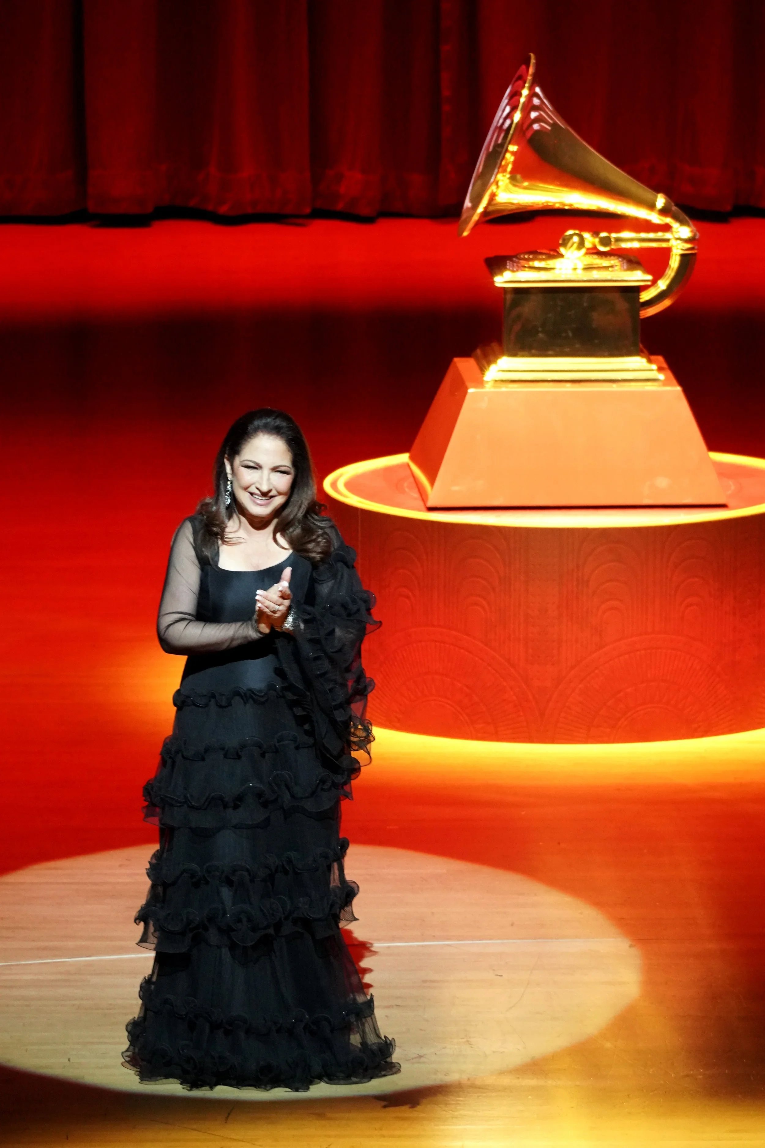 A woman in a black dress with ruffled layers standing on a stage with a large gramophone trophy displayed behind her.