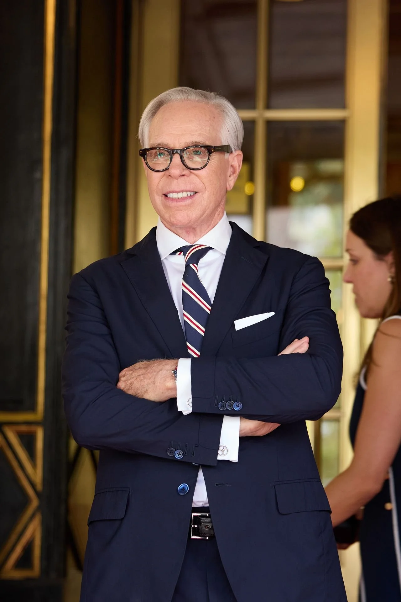 An elderly man with silver hair and glasses smiling, dressed in a dark suit, white shirt, and striped tie, standing with arms crossed in an indoor setting with a woman in the background.
