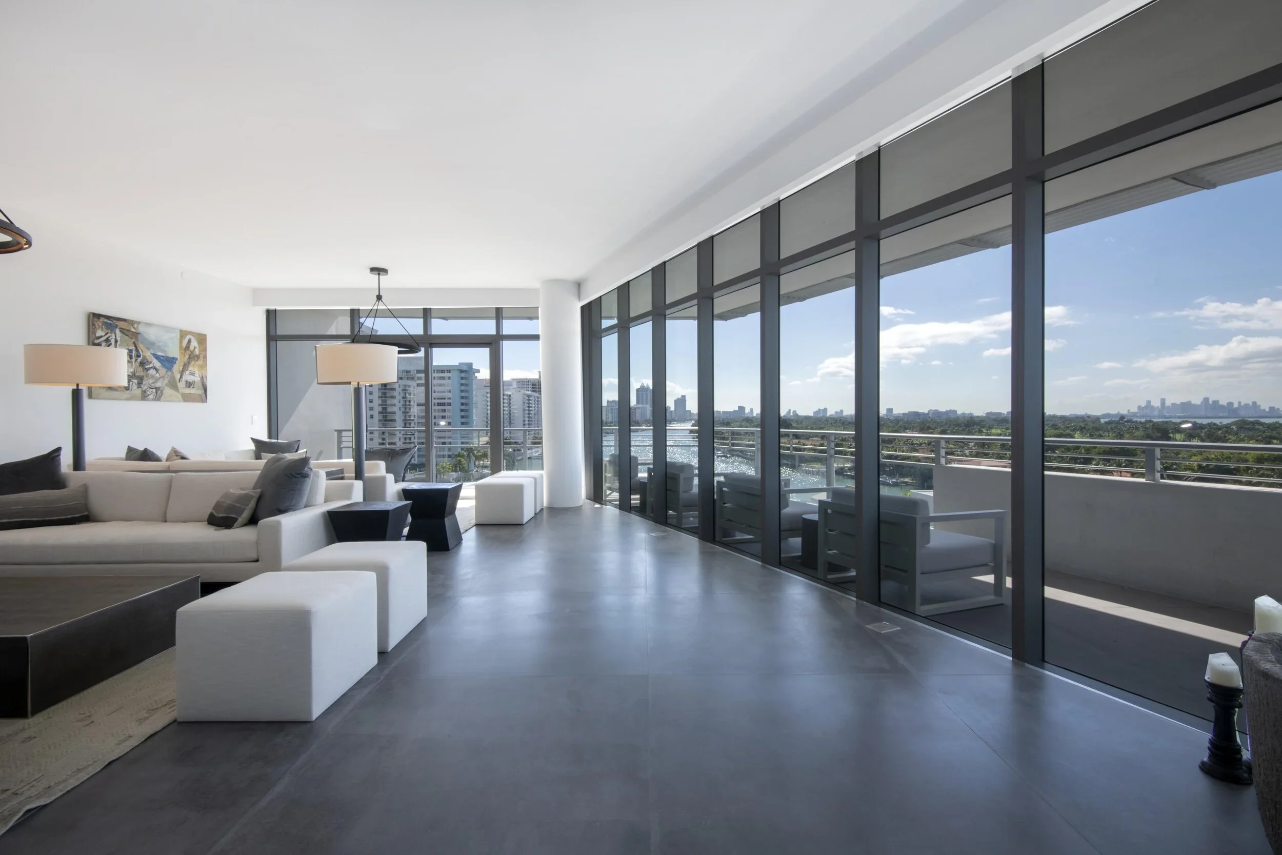 Modern living room with large floor-to-ceiling windows overlooking a cityscape and river, white sofas, black side tables, and contemporary artwork on white walls.
