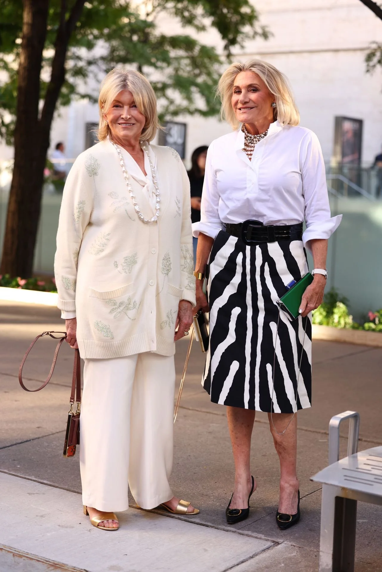 Two older women standing outdoors, dressed elegantly, one in a cream cardigan and pants with accessories, the other in a white shirt and a black-and-white zebra-striped skirt, with a pharmacy in the background.