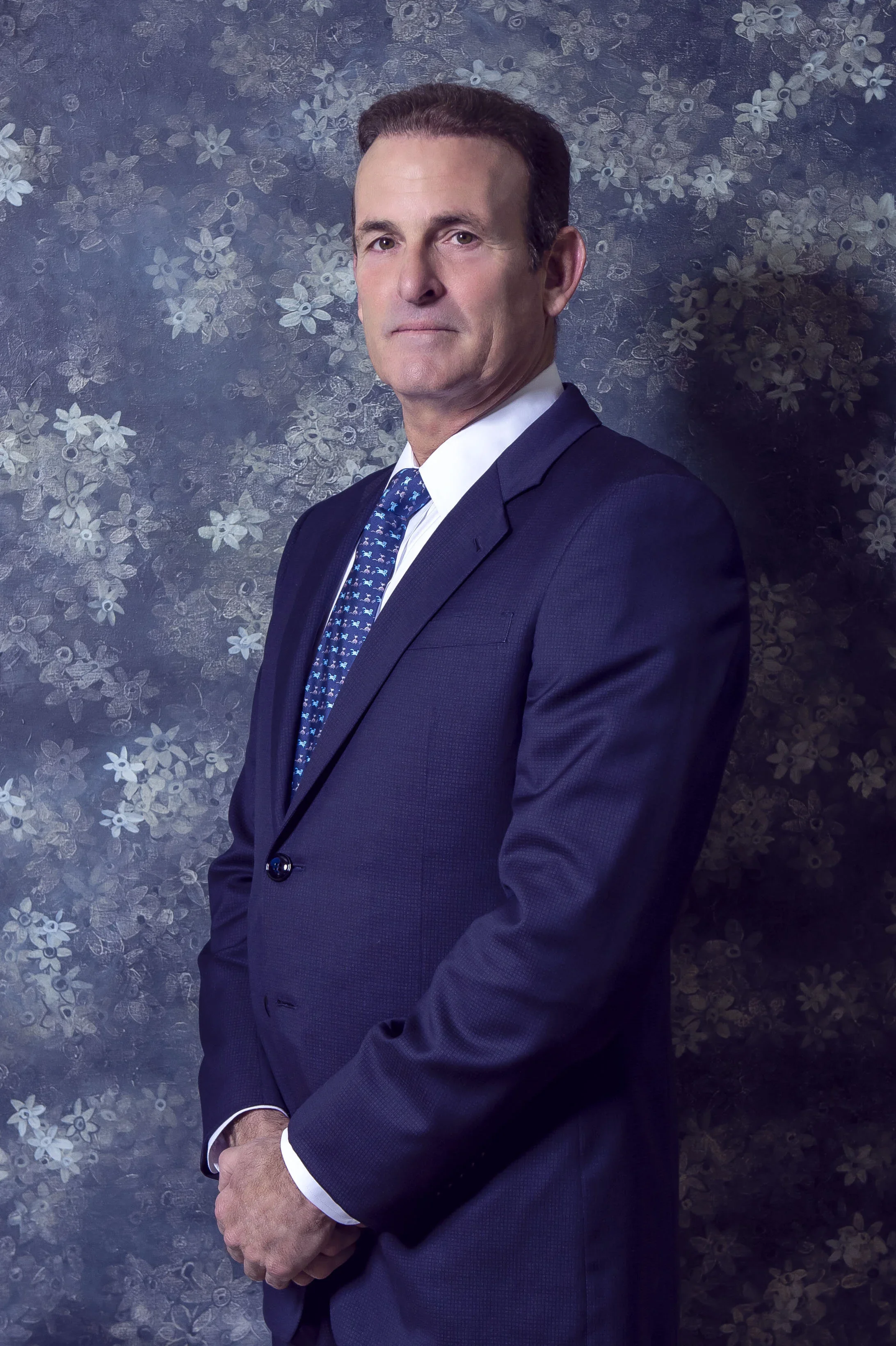 Portrait of a middle-aged man wearing a navy blue suit, white shirt, and a patterned blue tie, standing against a background with a floral design.