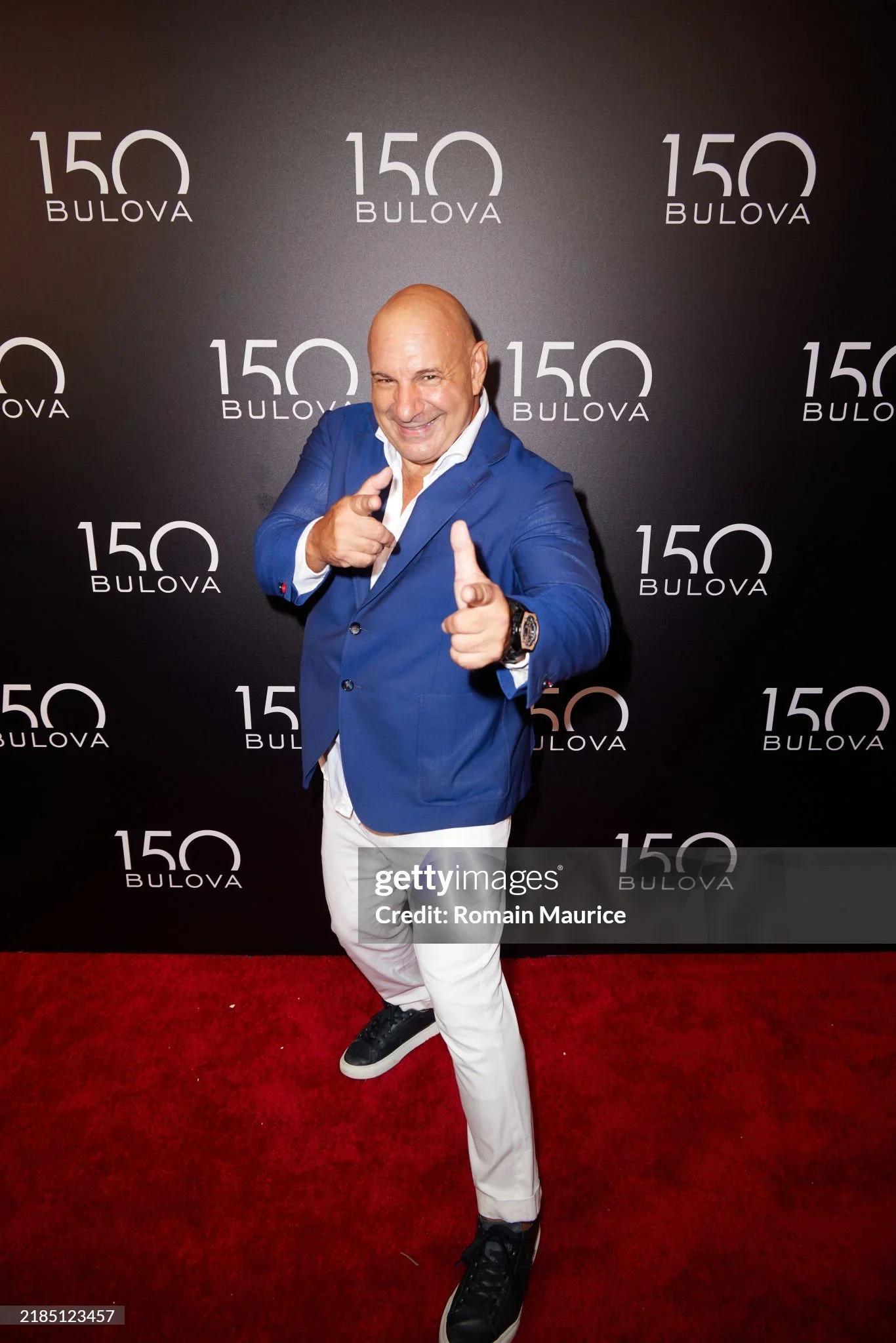 A man in a blue blazer pointing at the camera, smiling, standing on a red carpet in front of a black backdrop with the logo '150 Bulova' repeated across it.