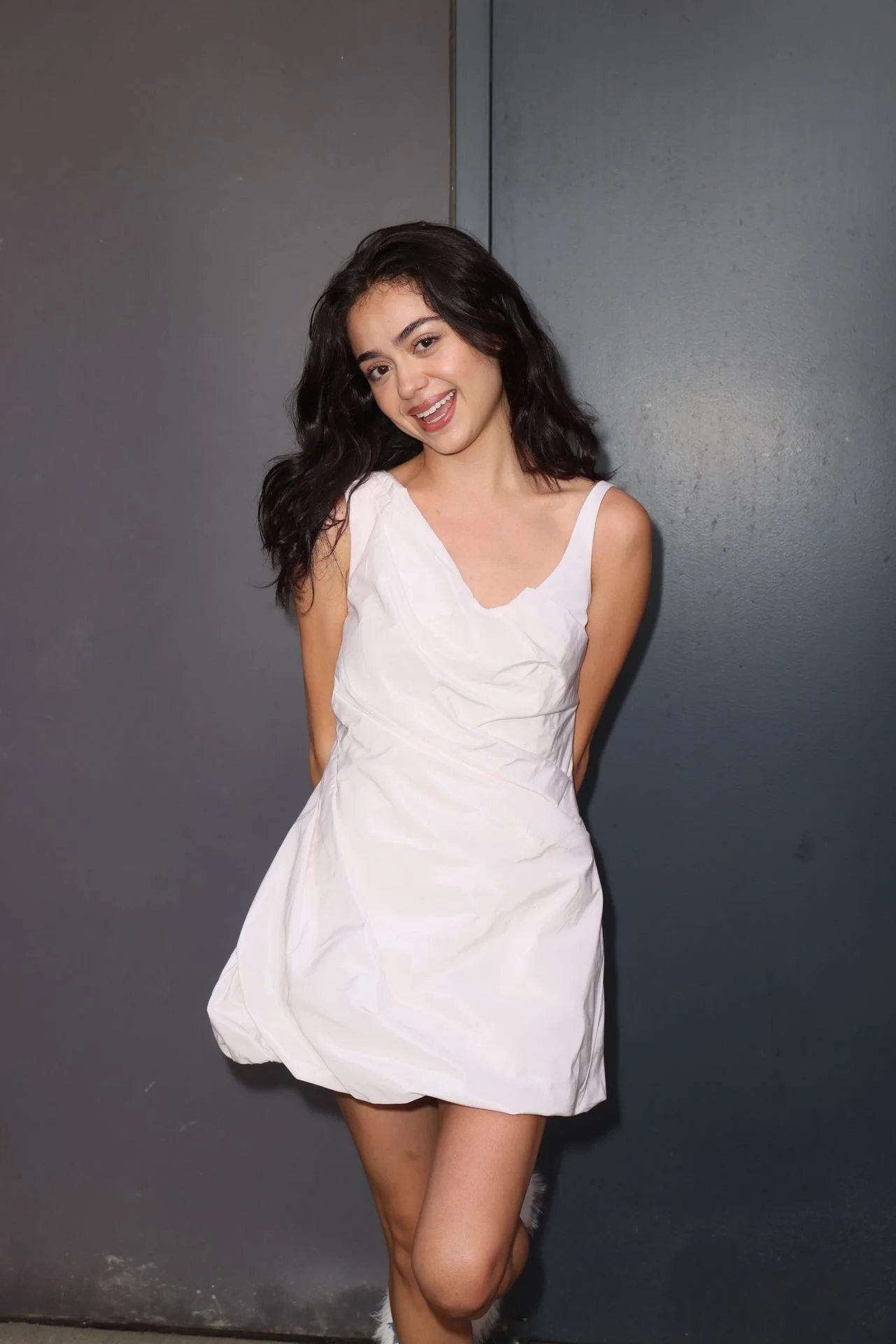 A young woman with dark, wavy hair styled in loose waves, smiling, wearing a sleeveless white dress with a slightly ruffled skirt, standing against a gray wall.