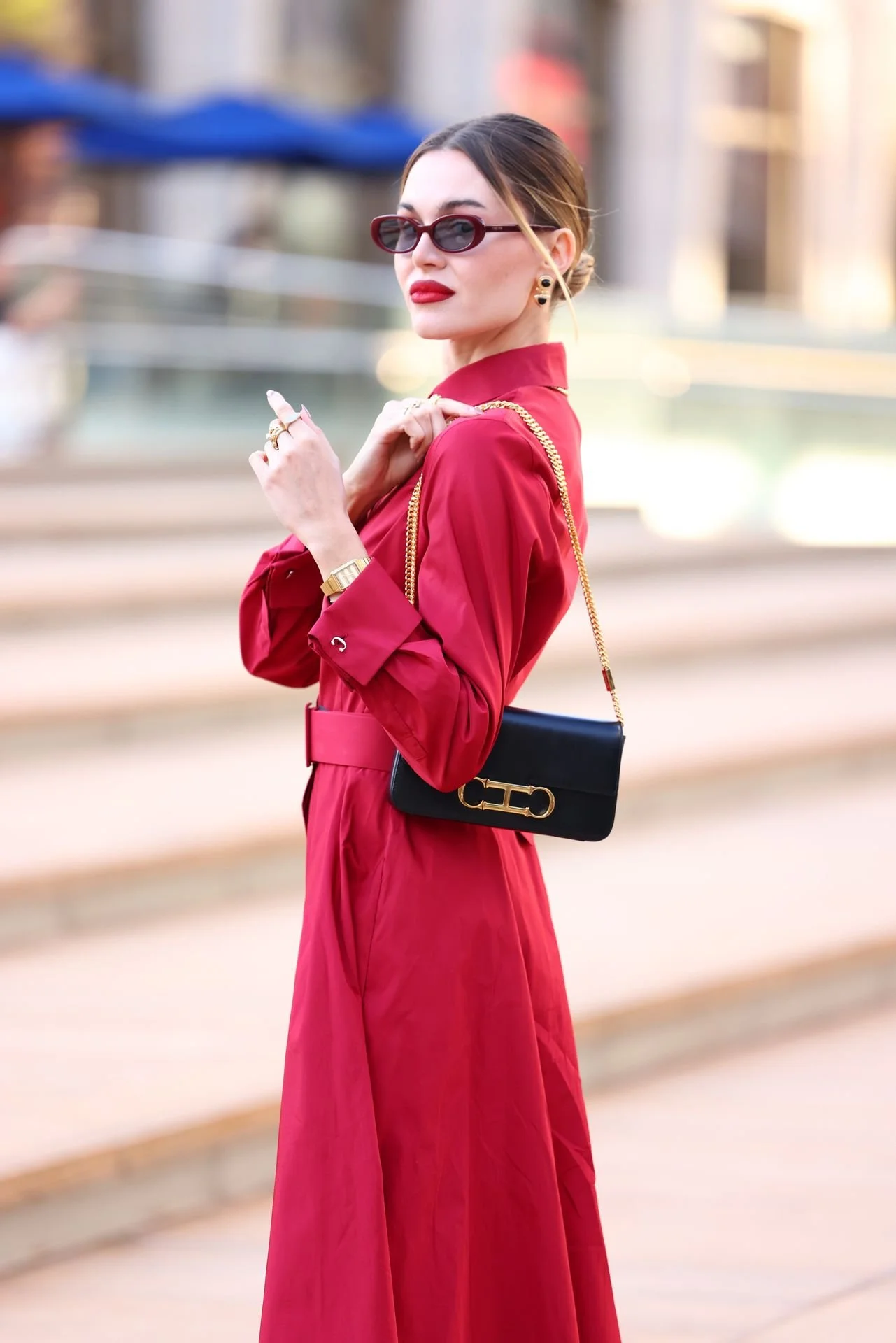 Stylish woman in red dress and sunglasses holding a black handbag with gold chain strap, standing outdoors.