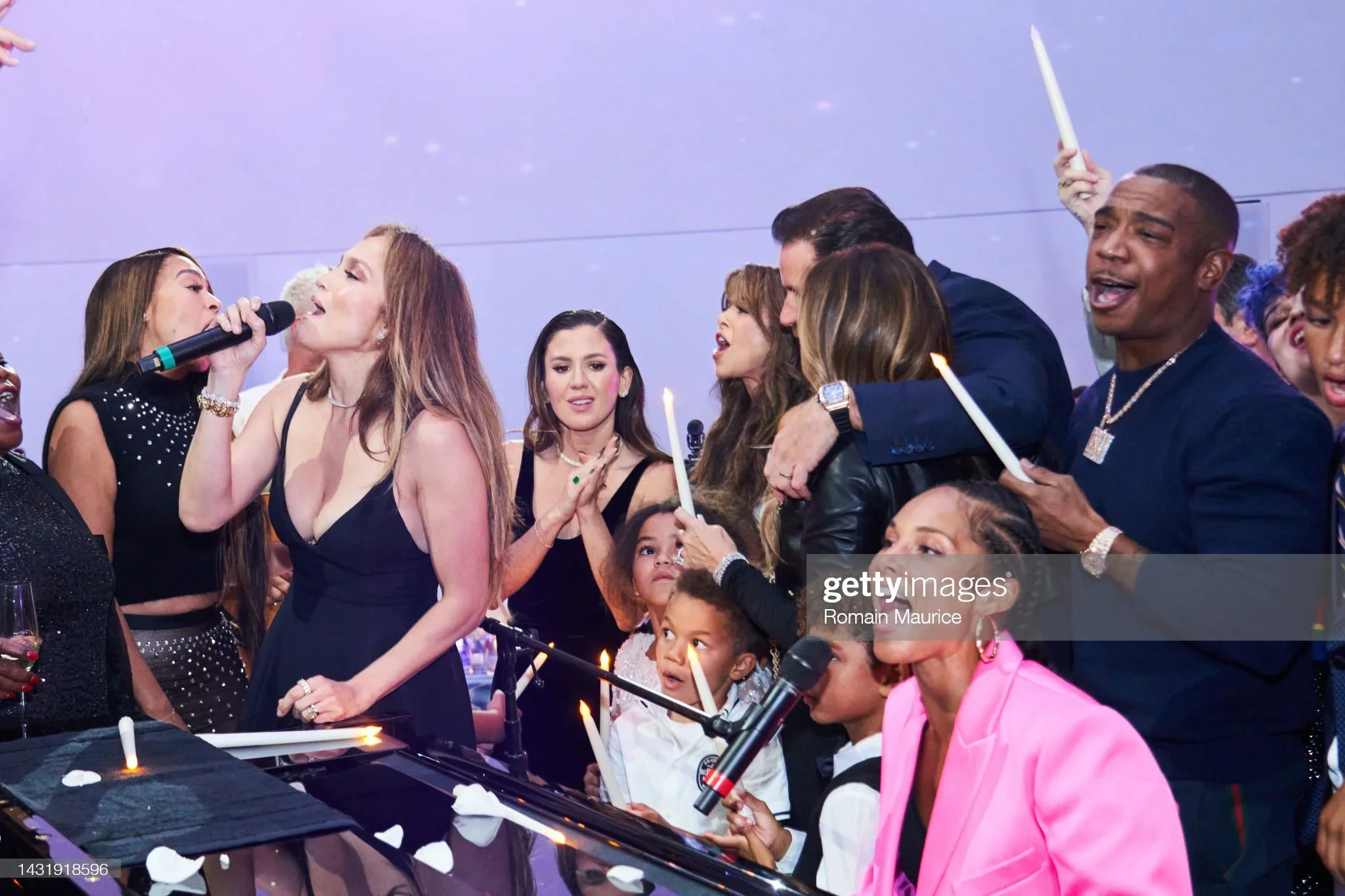 A group of people celebrating at a party with lit candles, some holding microphones, and a woman in a black dress singing or speaking into a microphone. The scene appears lively and festive.