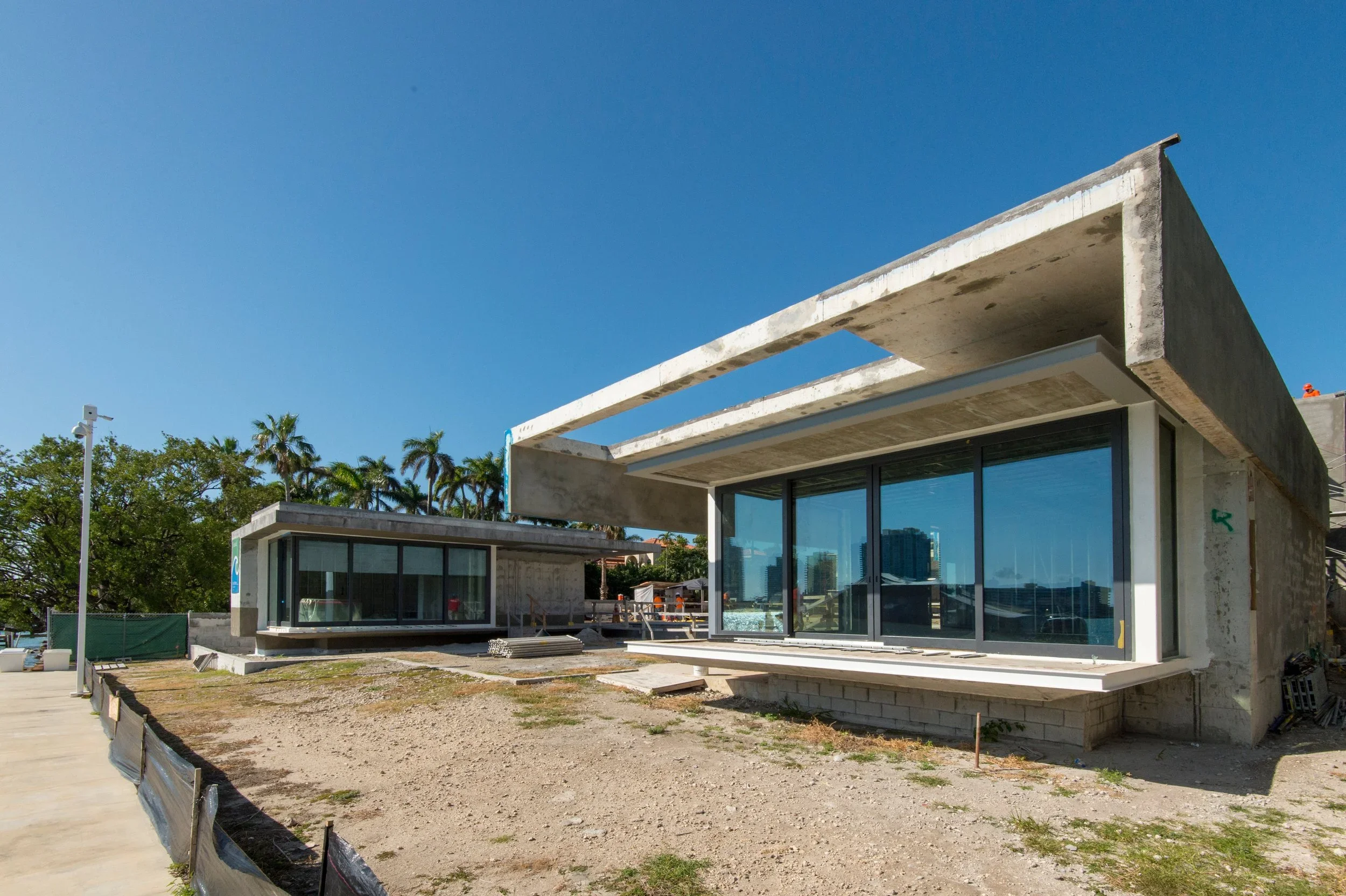 Modern house under construction with large glass windows, concrete structure, and a partially completed exterior. Construction materials and equipment are visible on site, surrounded by trees and clear blue sky.