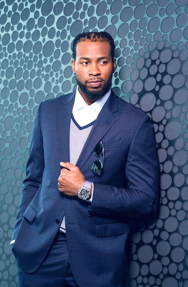 A man in a blue checkered suit standing against a circular patterned blue background, wearing sunglasses clipped to his jacket, with a wristwatch, and looking to the side.