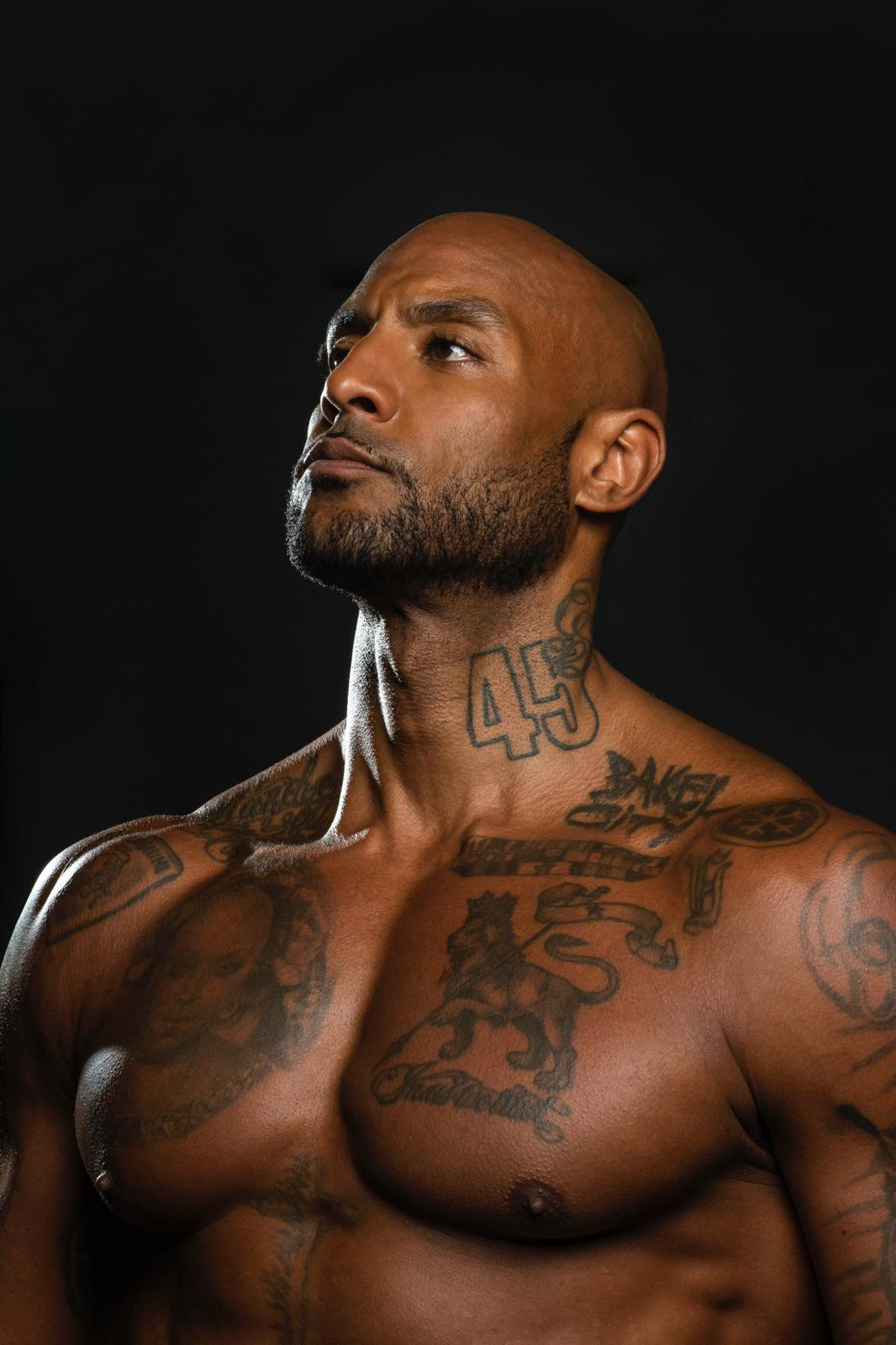 A shirtless man with a bald head and numerous tattoos on his chest, neck, and shoulders, looking slightly upward with a serious expression against a black background.