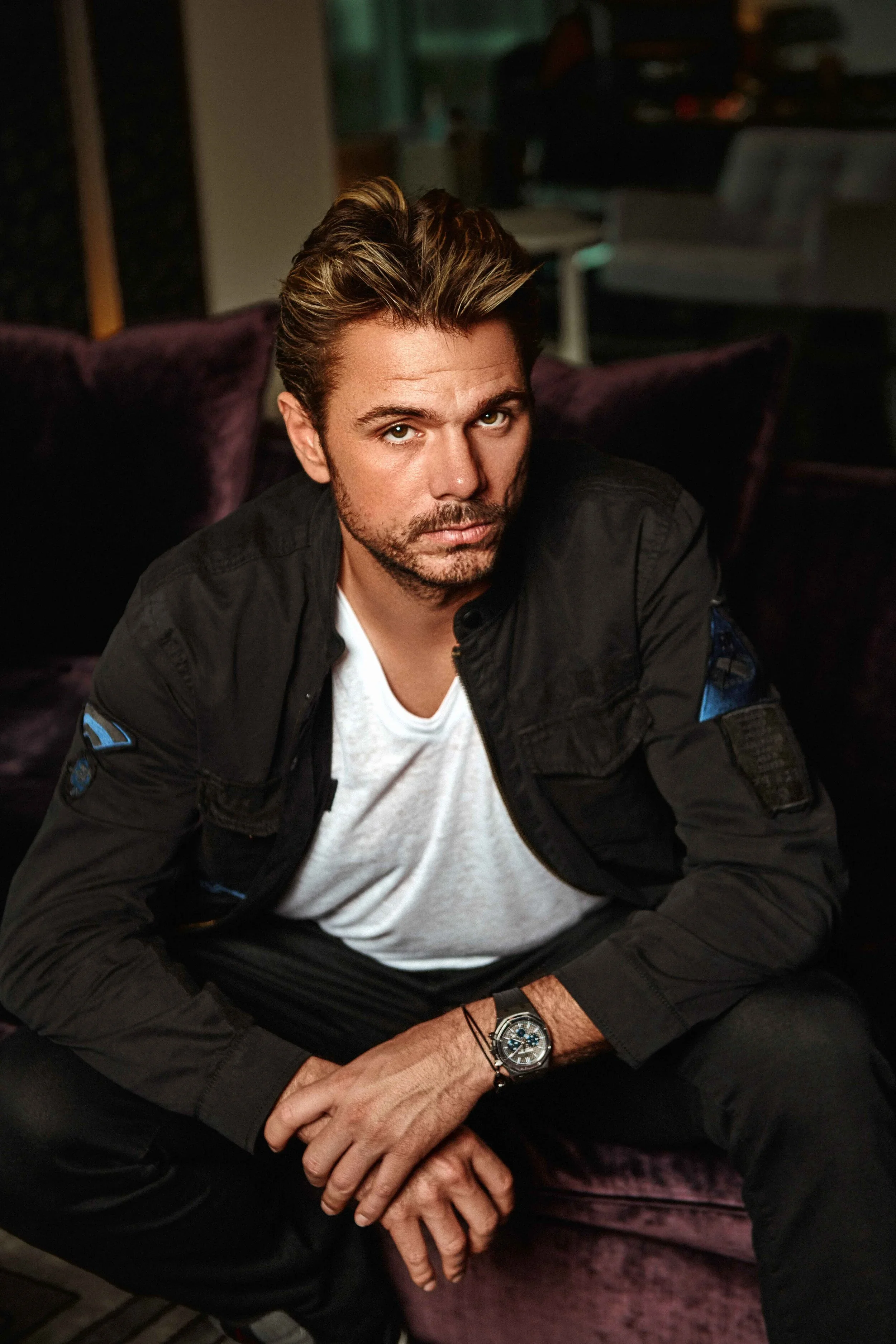 A man sitting on a purple couch, looking directly at the camera with a serious expression, wearing a black jacket, white t-shirt, and a wristwatch, in a dimly lit room.