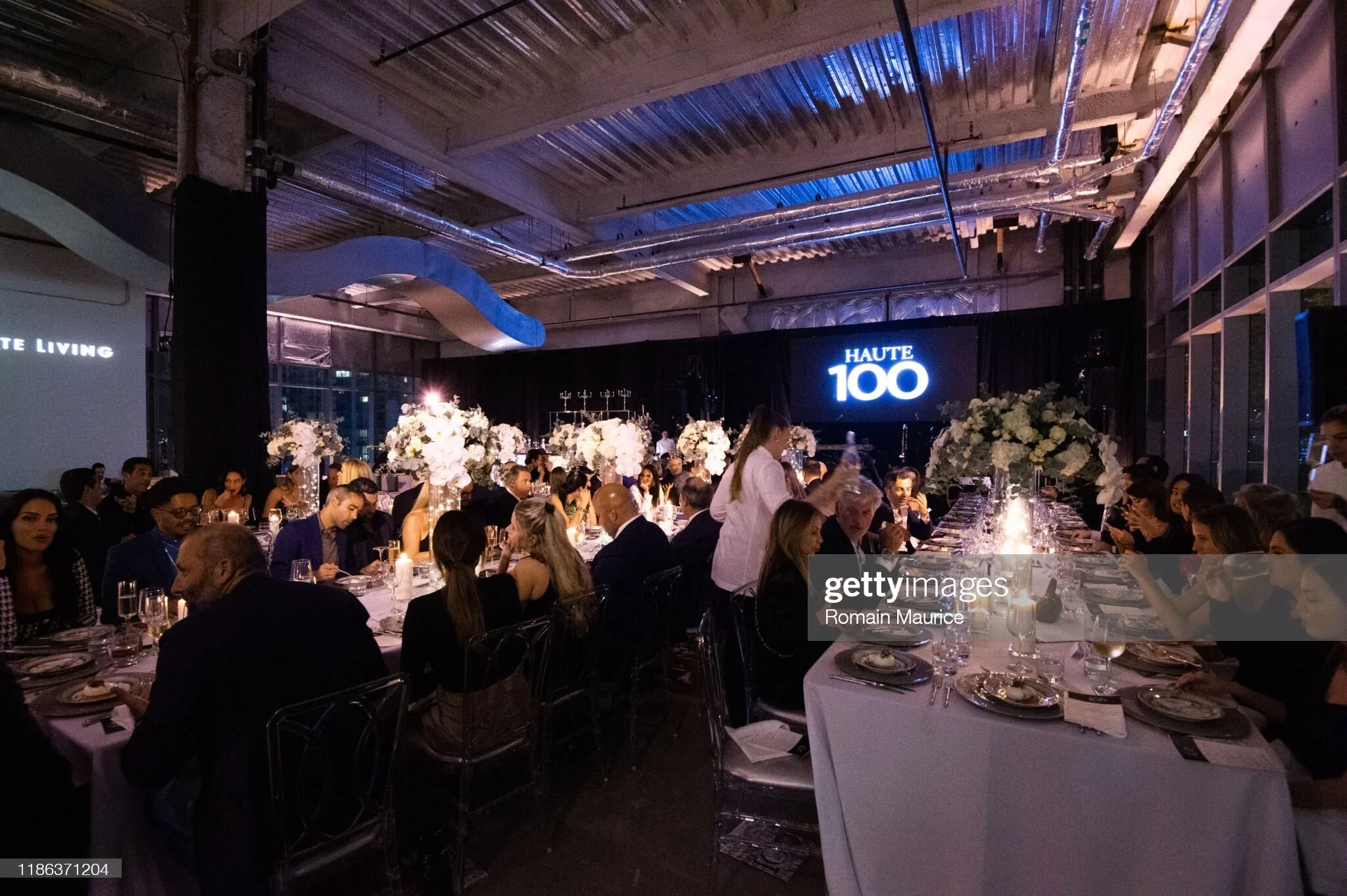 A large indoor banquet hall filled with round tables decorated with white floral centerpieces and candles. There are many people dressed in formal attire seated at the tables. In the background, a screen displays the words 'Haute 100'. The ceiling ha