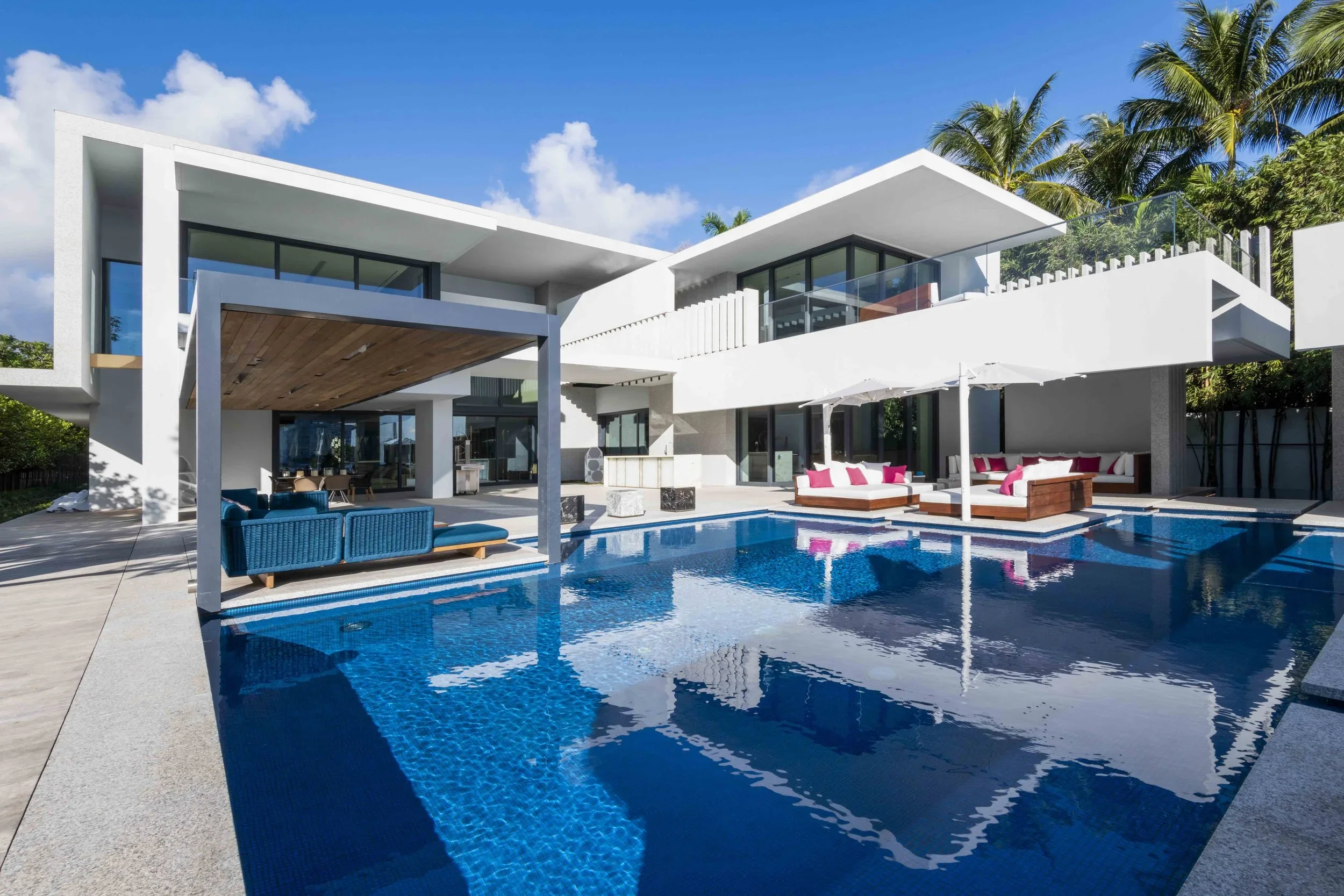 Modern white house with multiple levels and large glass windows, outdoor patio area with seating, lounge chairs, and umbrellas, swimming pool, and tropical palm trees under a blue sky.
