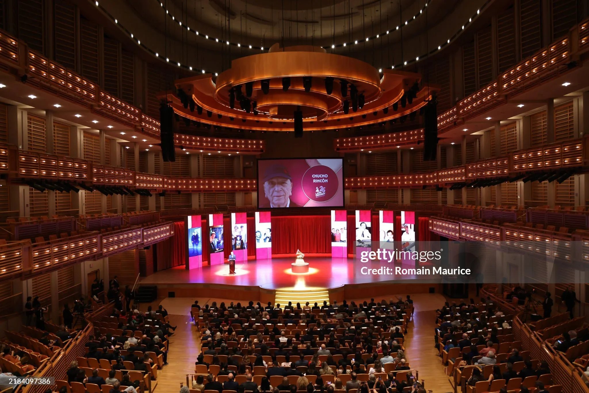 Inside a large auditorium with a stage and a big screen displaying a person and the text 'Chucho Rincón.' The auditorium has multiple levels with wooden balconies and is filled with seated audience members.