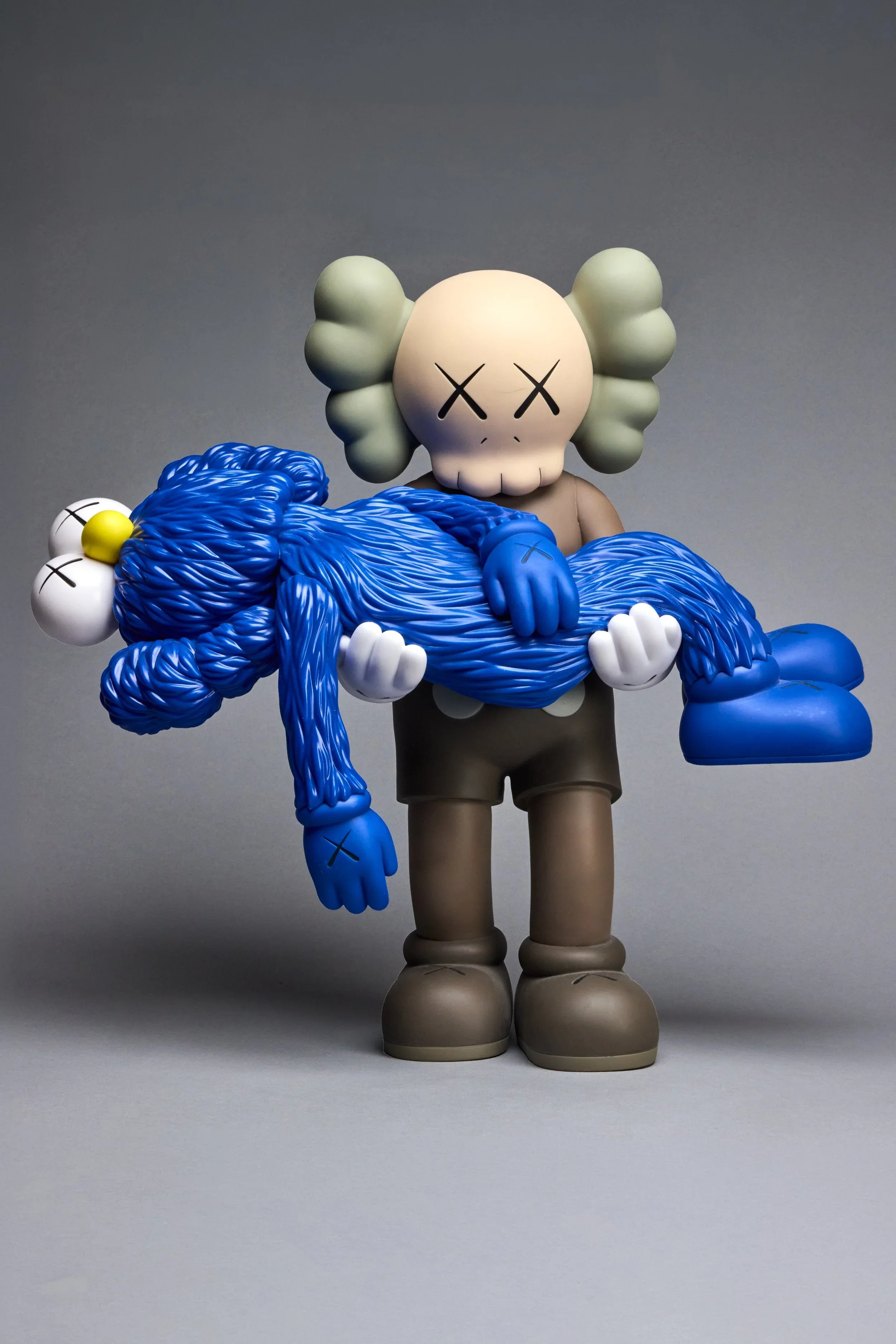 A cartoon-style figure with a skull face and cloud-like hair, wearing beige shoes and gray shorts, holding a large blue yarn-like object with two arms and two hands, in front of a plain gray background.