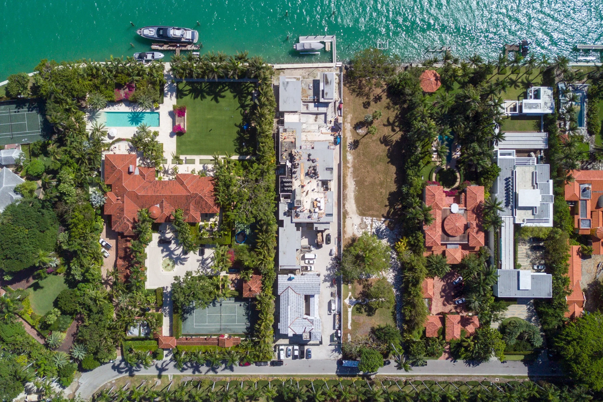 An aerial view of a coastal residential area with a waterway and several boats docked, surrounded by lush green trees and houses with red-tile roofs.
