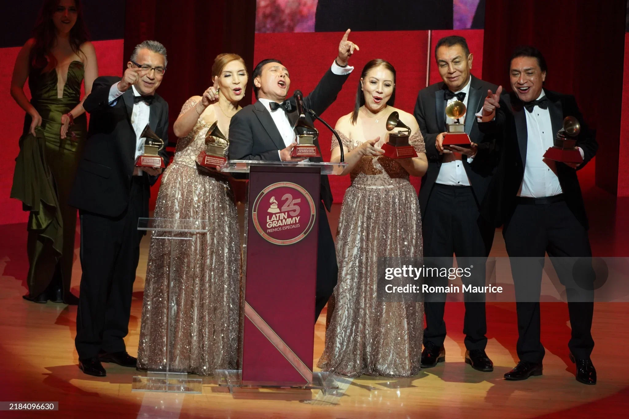 A group of seven well-dressed people celebrating at an award ceremony, holding Grammy trophies. The men are in tuxedos, and the women are in elegant, gold-sequined dresses, standing behind a podium with a red logo that reads '25 Latin Grammy Premios 