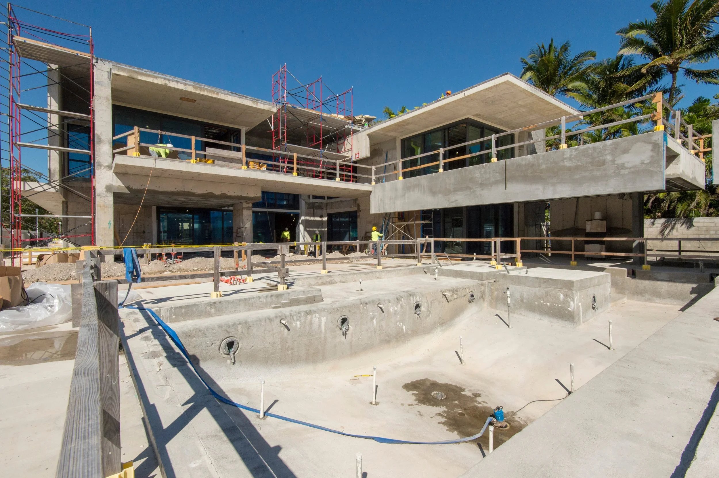A modern house under construction with concrete structure, scaffolding, and construction workers wearing yellow helmets, with palm trees and a clear blue sky in the background.