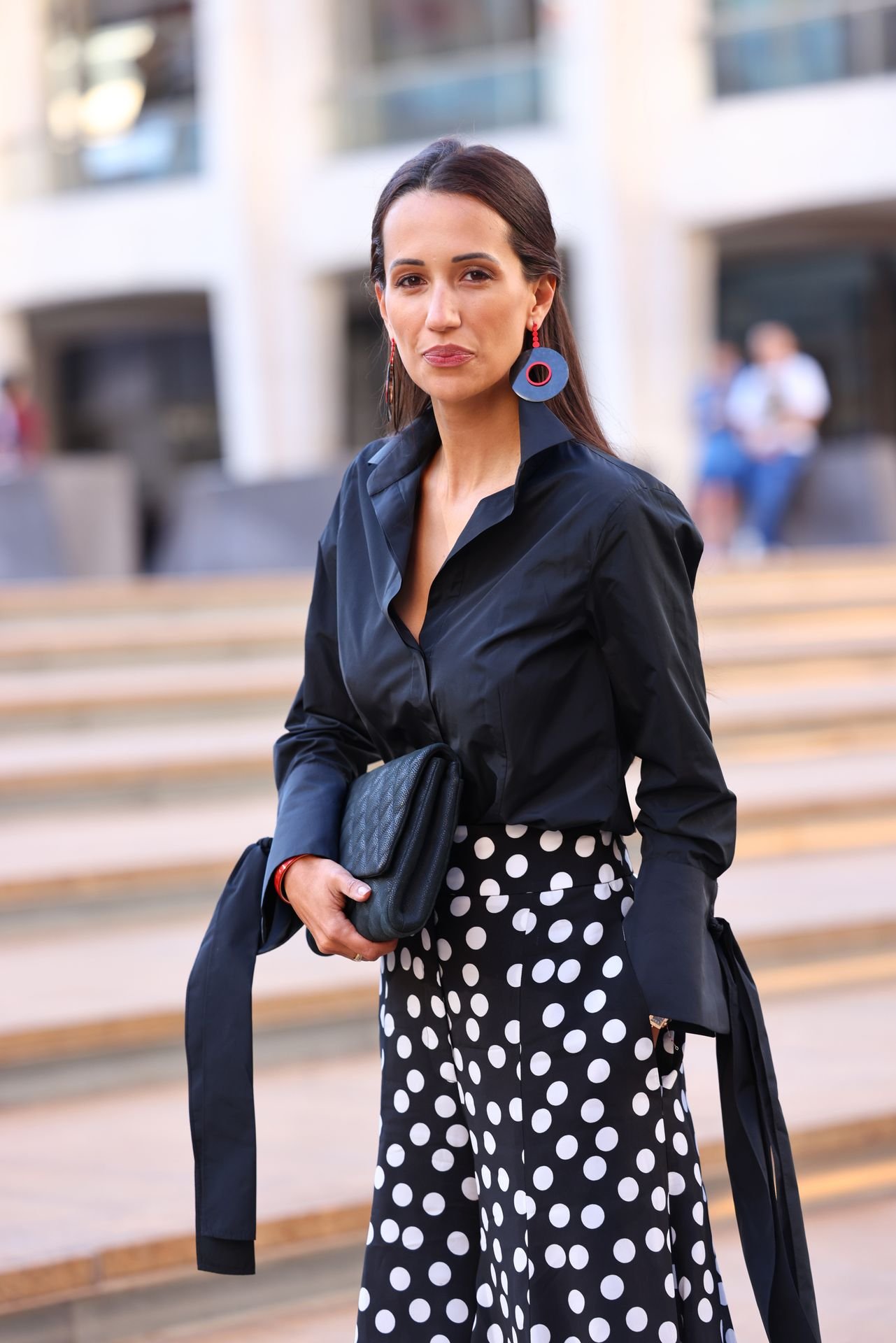 A woman with dark brown hair wearing a black button-up shirt, polka dot skirt, and red earrings, holding a black clutch purse, standing outdoors with a modern building in the background.