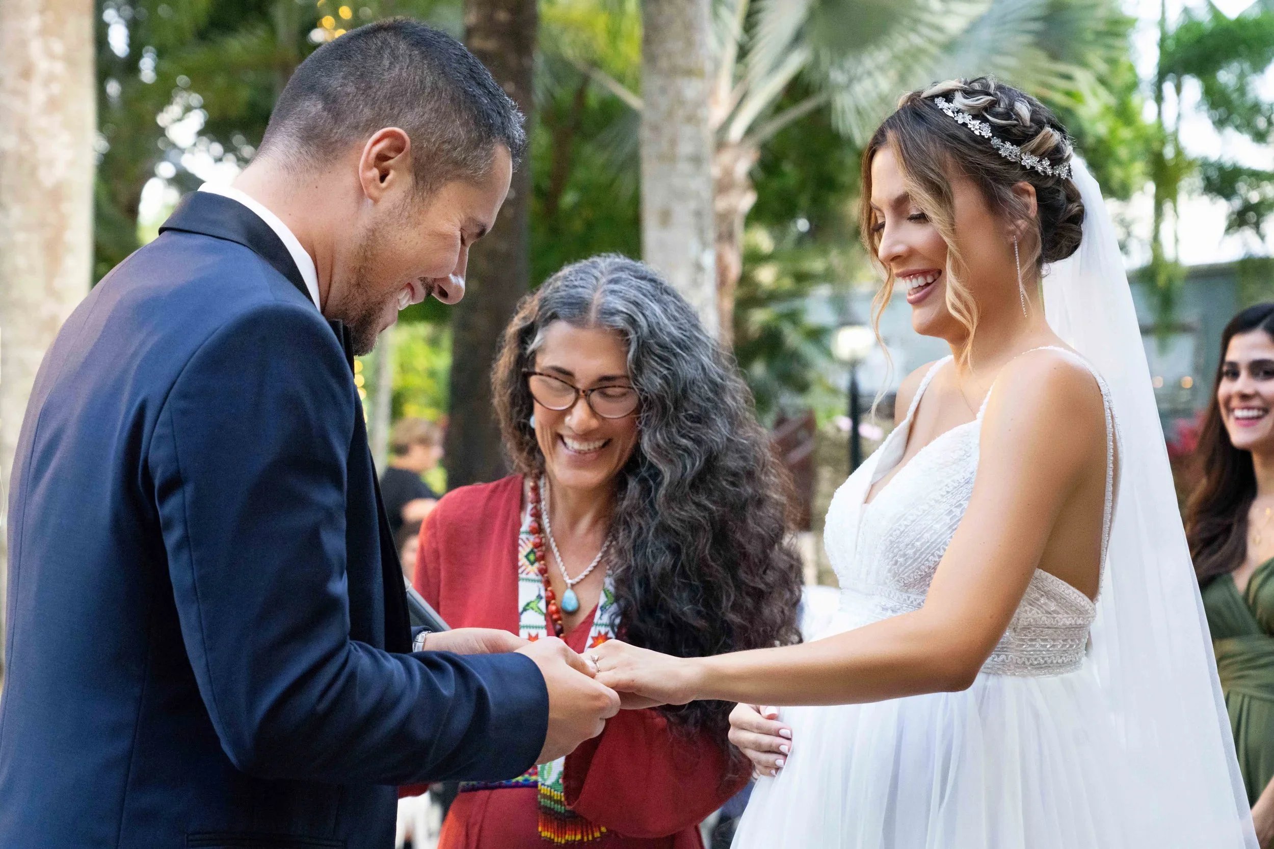 A couple exchange vows during a wedding ceremony outdoors, with a woman officiant and a bridesmaid visible, surrounded by greenery and trees.