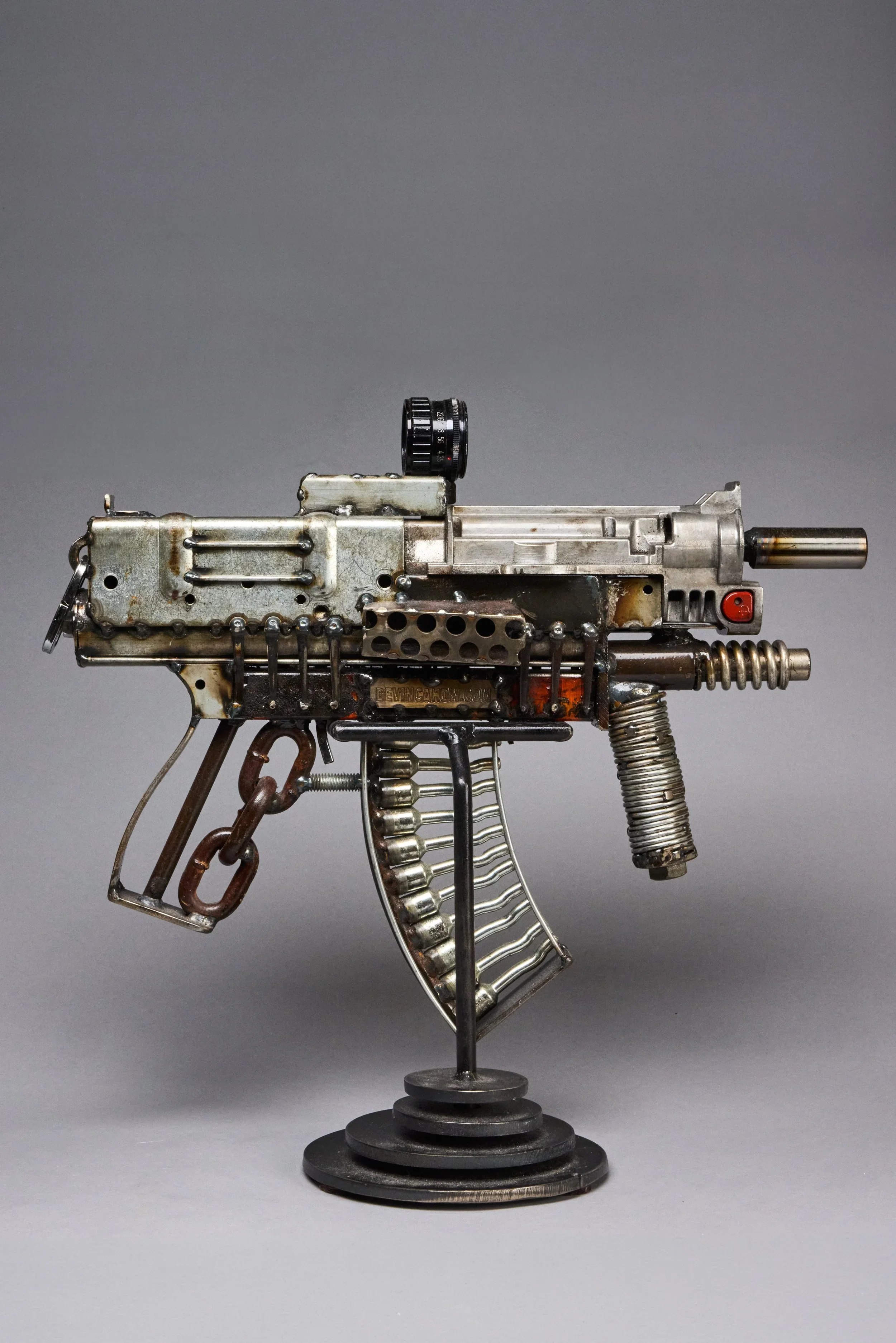 A sculpture resembling a firearm made from metallic parts, chains, and mechanical components, standing on a circular base.