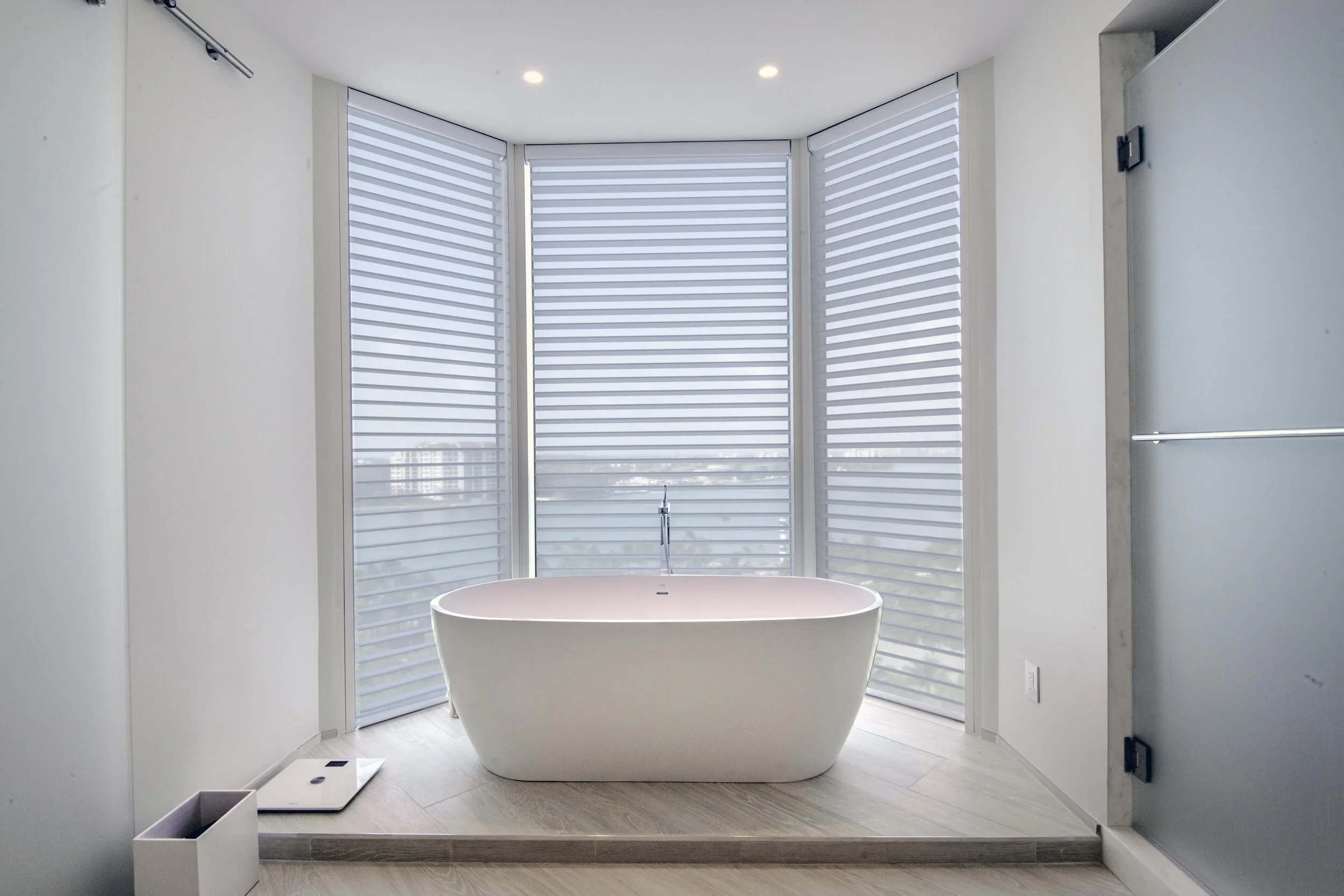 Modern bathroom with a freestanding bathtub in front of large multi-pane windows with white horizontal blinds, a digital scale on the floor, and a small white container on the ledge.
