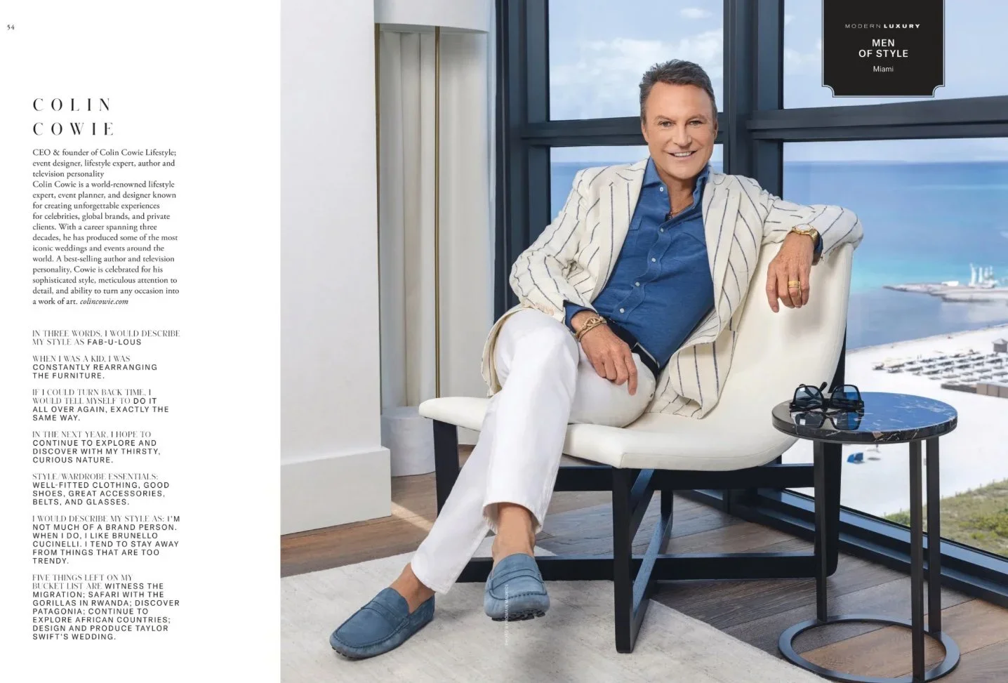 A man dressed in a blue shirt, white pants, and a light-colored striped blazer sitting in a modern apartment with large windows overlooking the ocean. He is smiling, wearing rings and bracelets, and has sunglasses on a small black marble table beside