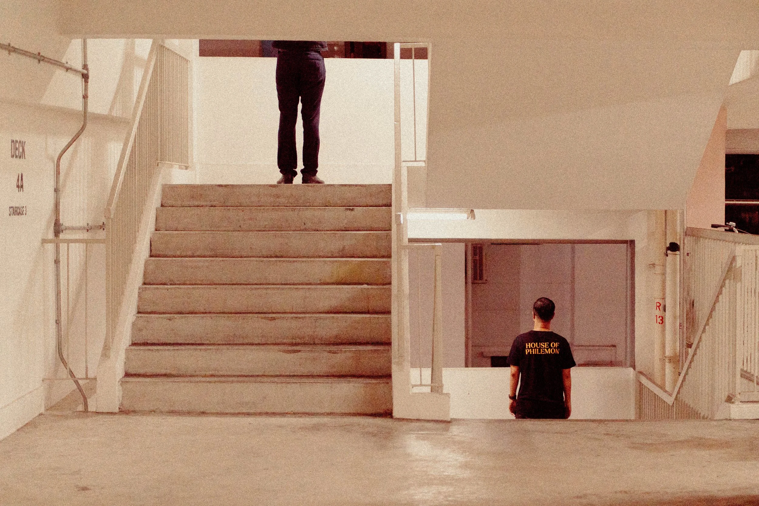 Two people standing in an indoor parking garage. One person is on the upper level by the staircase, and the other is on the lower level near the bottom of the stairs. The person on the lower level is wearing a black T-shirt with yellow text that read