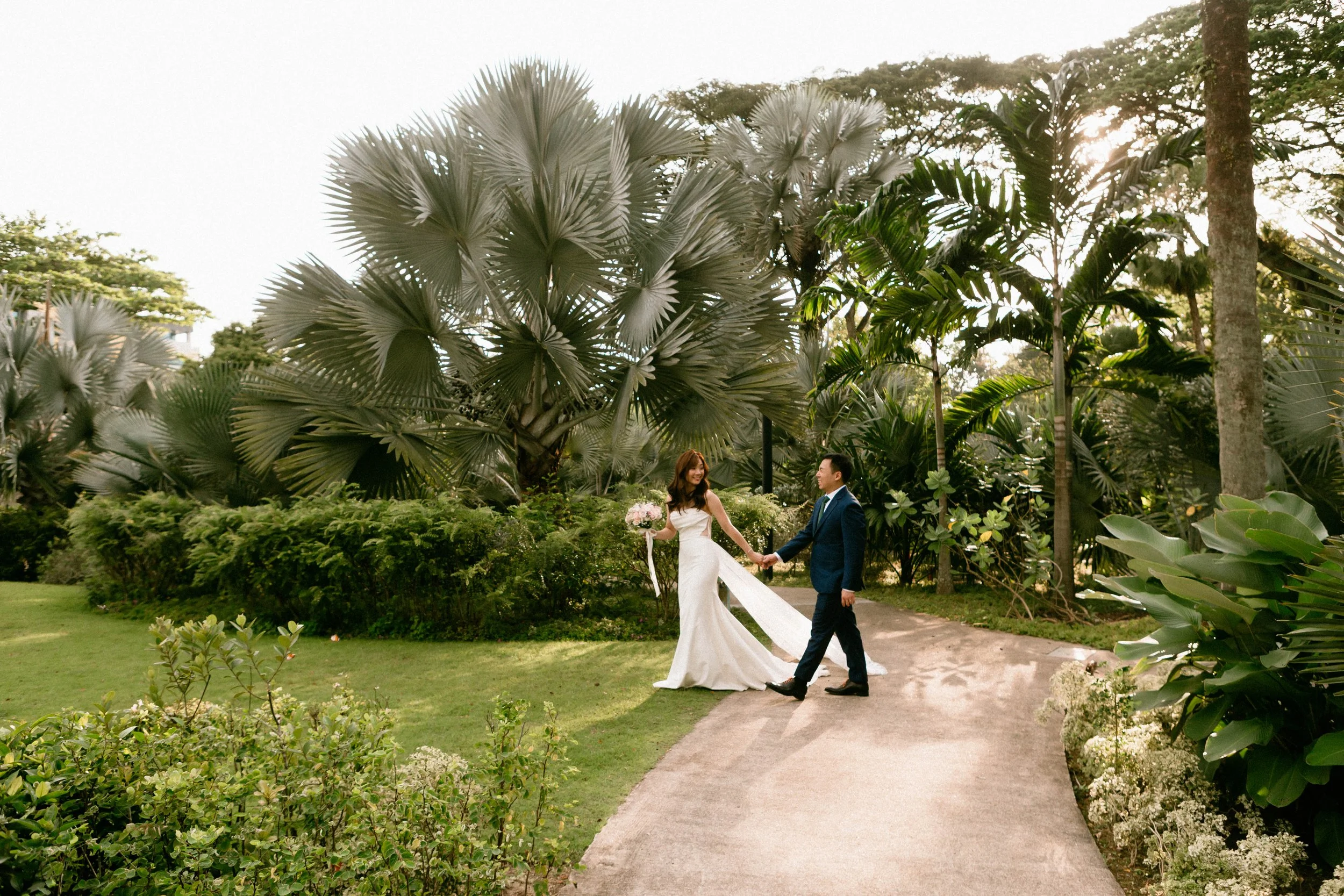 A bride and groom holding hands and walking on a garden path surrounded by lush greenery and large tropical plants, with sunlight filtering through the trees.